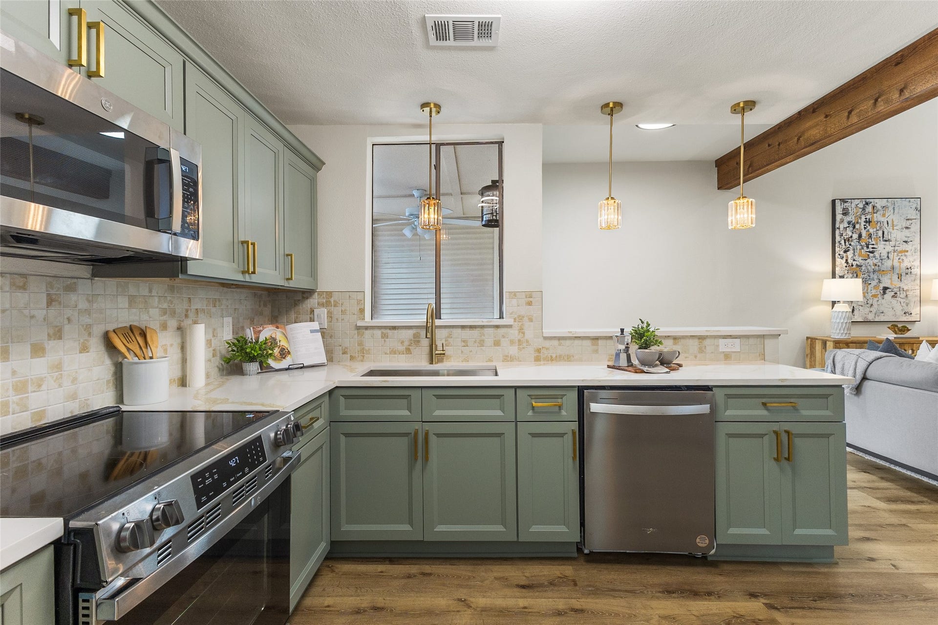 Beautifully updated kitchen features sage-green cabinetry, gold hardware, sleek white countertops, stainless steel appliances, decorative tile backsplash, and elegant pendant lighting overlooking the living area.