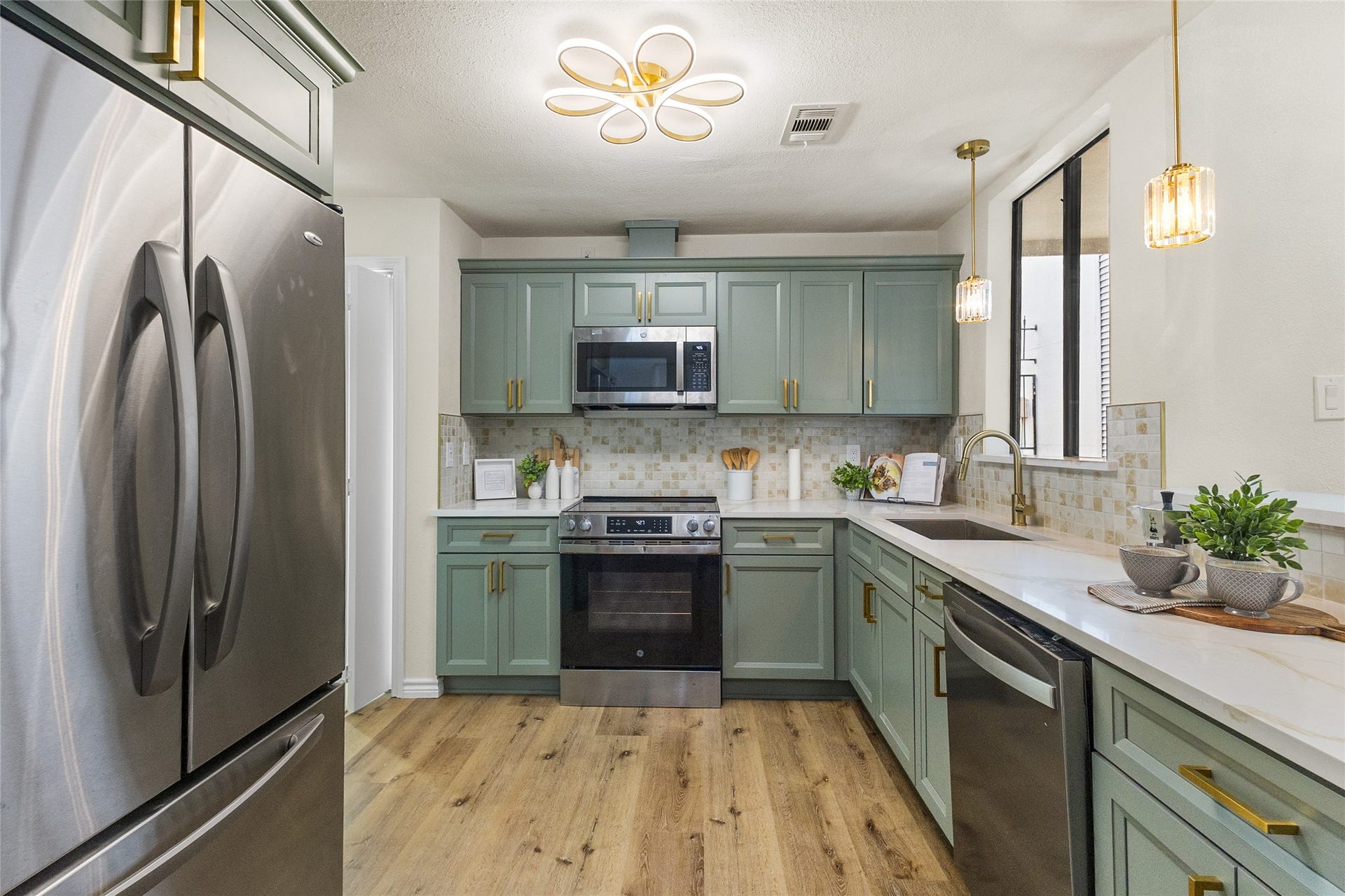 Your kitchen features sage-green cabinetry with elegant gold hardware, sleek white countertops, stainless steel appliances, a decorative tile backsplash, modern lighting fixtures, and beautiful wood-look flooring.