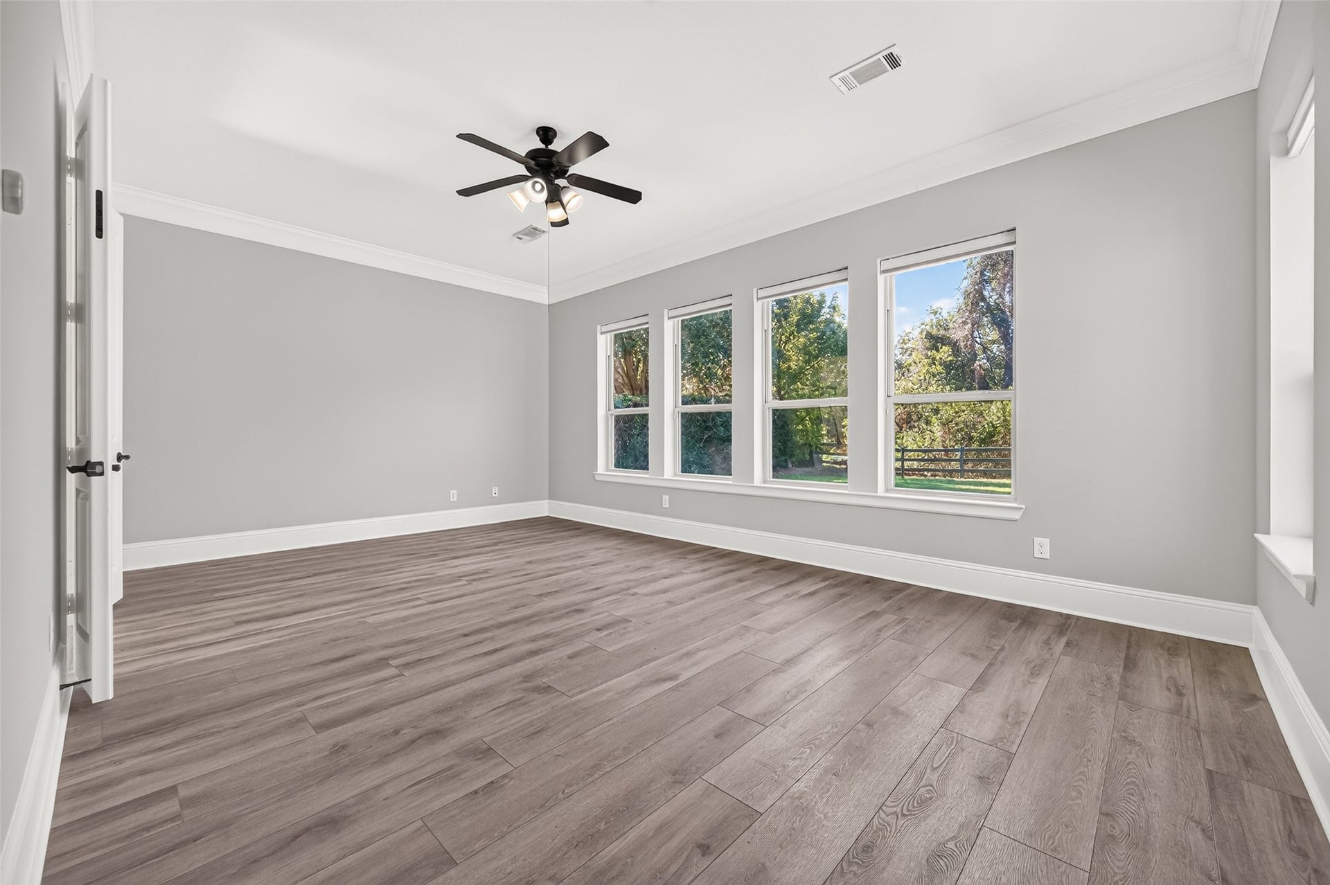 5119 Water Oak Crescent, Fulshear, TX 77441 Serene primary suite retreat! This spacious bedroom boasts stylish, modern wood-look flooring, elegant crown molding, a classic ceiling fan, and a wall of large windows overlooking the lush, private backyard space.
