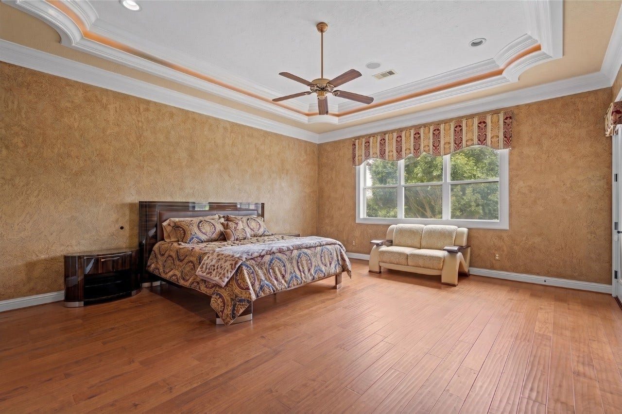 Spacious secondary suite upstairs showcasing an ornate illuminated tray ceiling, rich hardwood flooring, and large windows with serene treetop views.