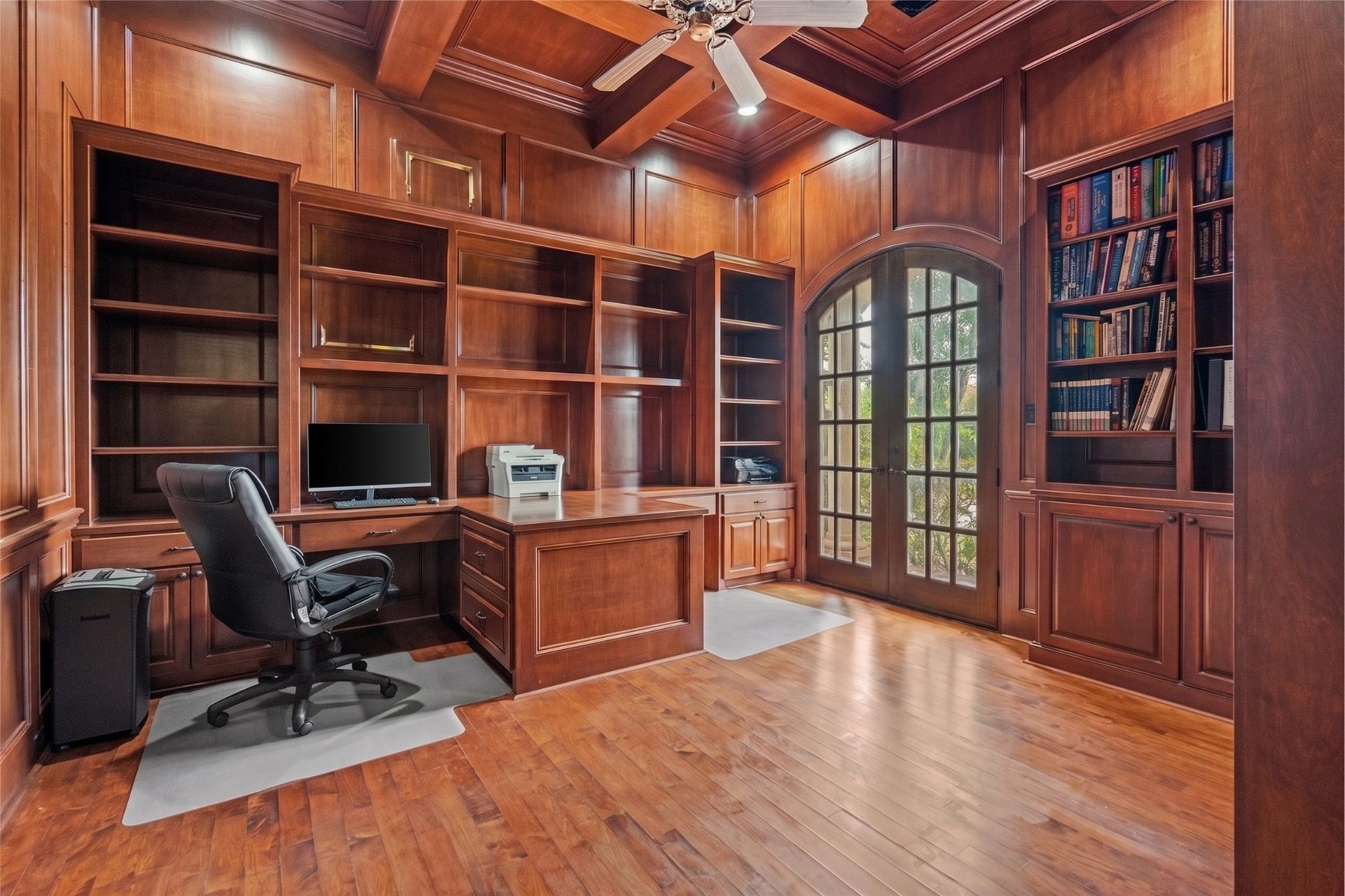 Luxurious executive home office featuring rich wood panelling, custom built-ins, a coffered ceiling, hardwood floors, and elegant arched French doors.