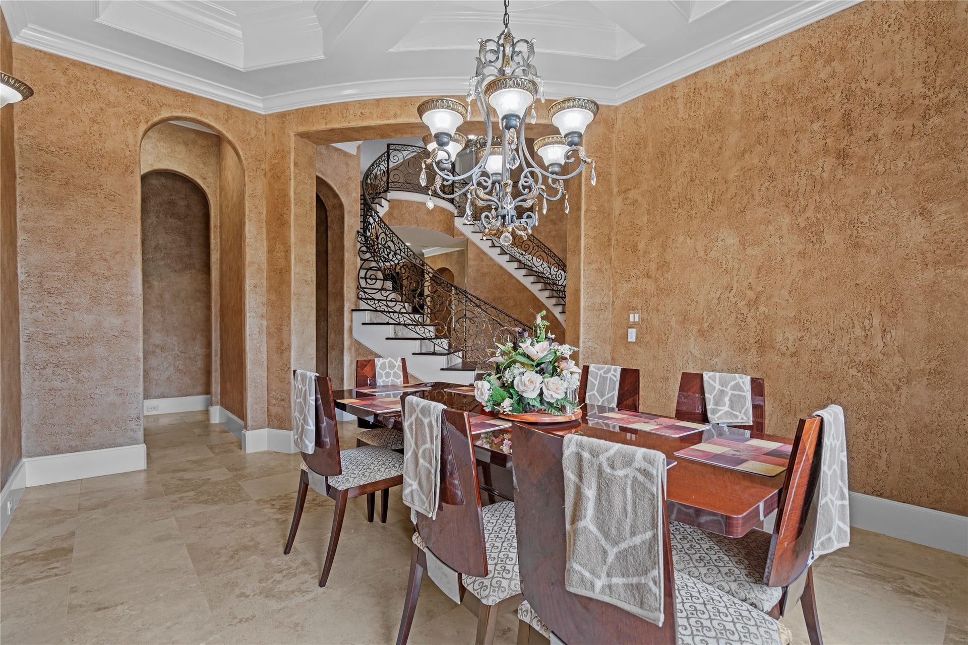 Formal dining room featuring a sculpted tray ceiling, ornate chandelier, and graceful architectural details with a striking curved double staircase beyond.