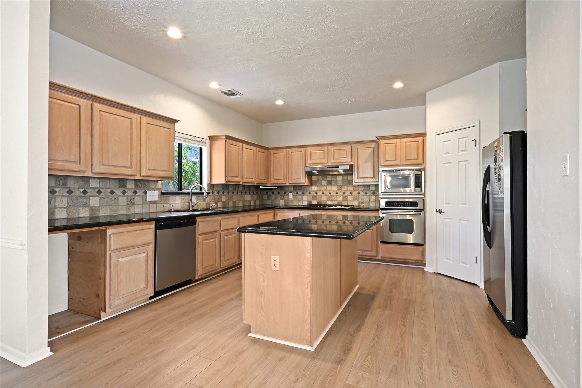 2607 Sailboat Drive, Houston, TX 77058 Kitchen accented with light wood cabinetry, sleek black countertops, and a central island, complemented by stainless steel appliances and a stylish tile backsplash.