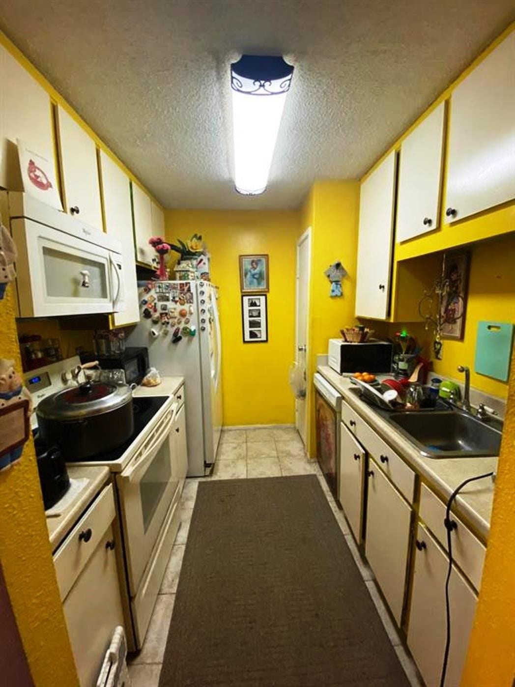 10049 Westpark Drive, #136, Houston, TX 77042 This cozy, narrow kitchen features bright yellow walls and white cabinetry, equipped with essential appliances like a stove, microwave, and refrigerator. It has overhead lighting, tile flooring, and ample storage space, creating a warm and functional cooking area.
