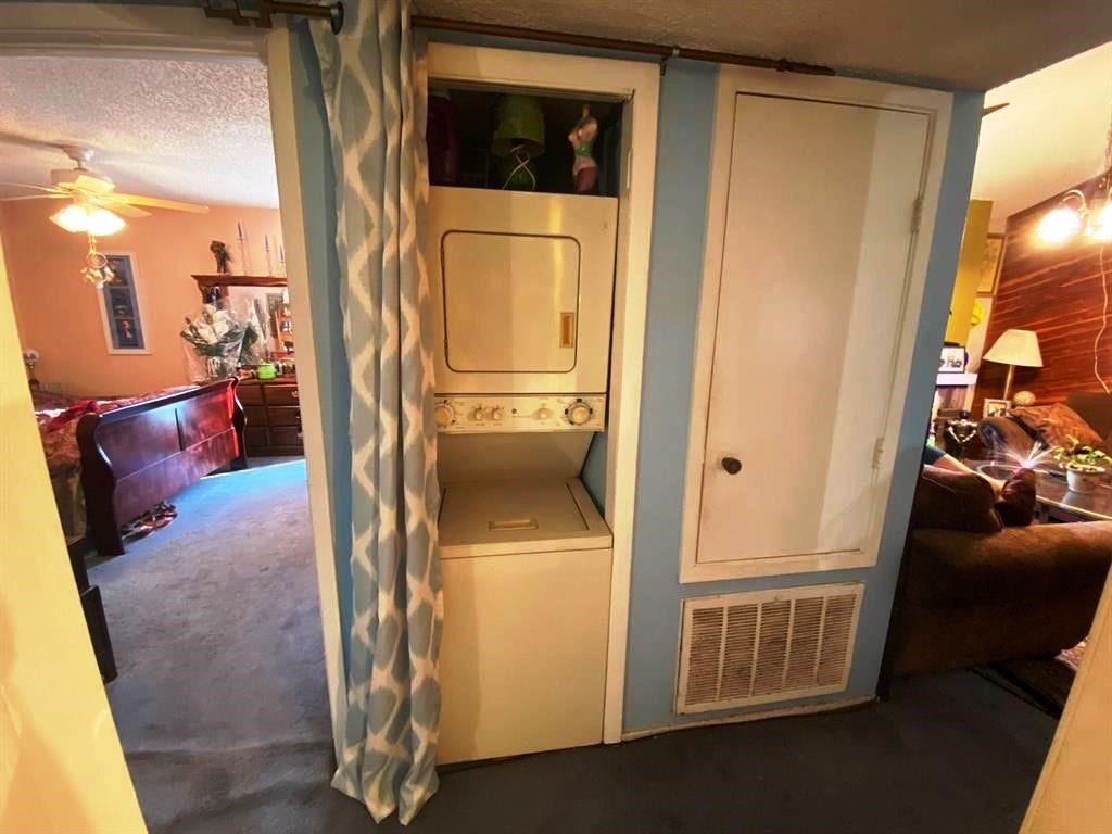 10049 Westpark Drive, #136, Houston, TX 77042 This photo shows a compact laundry area with a stacked washer and dryer unit, separated by a curtain. It is situated between a cozy living space and a bedroom, featuring a warm color palette and carpeted flooring.