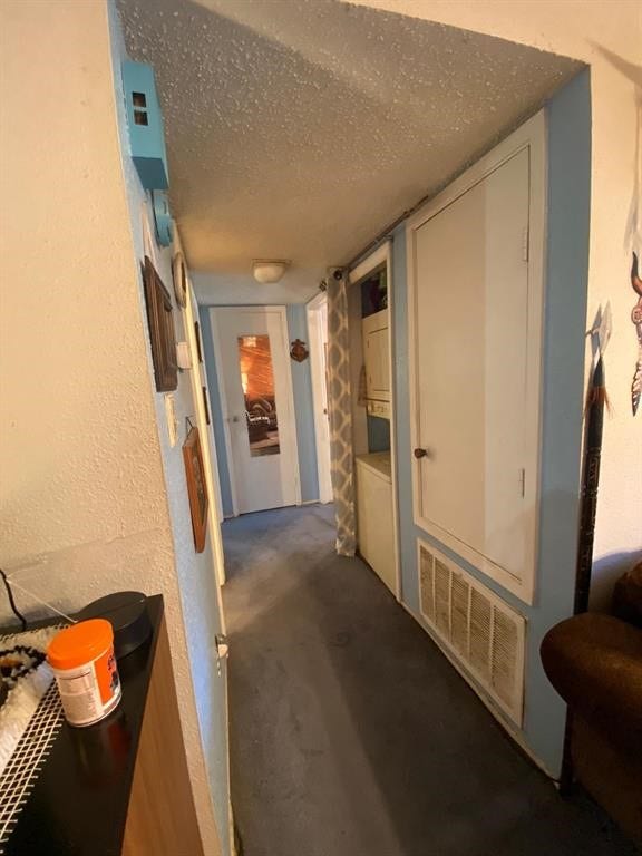10049 Westpark Drive, #136, Houston, TX 77042 A narrow hallway with textured ceilings, leads to a room with a partially open door. It features a built-in cabinet, a small utility area with a stacked washer and dryer, and a carpeted floor.