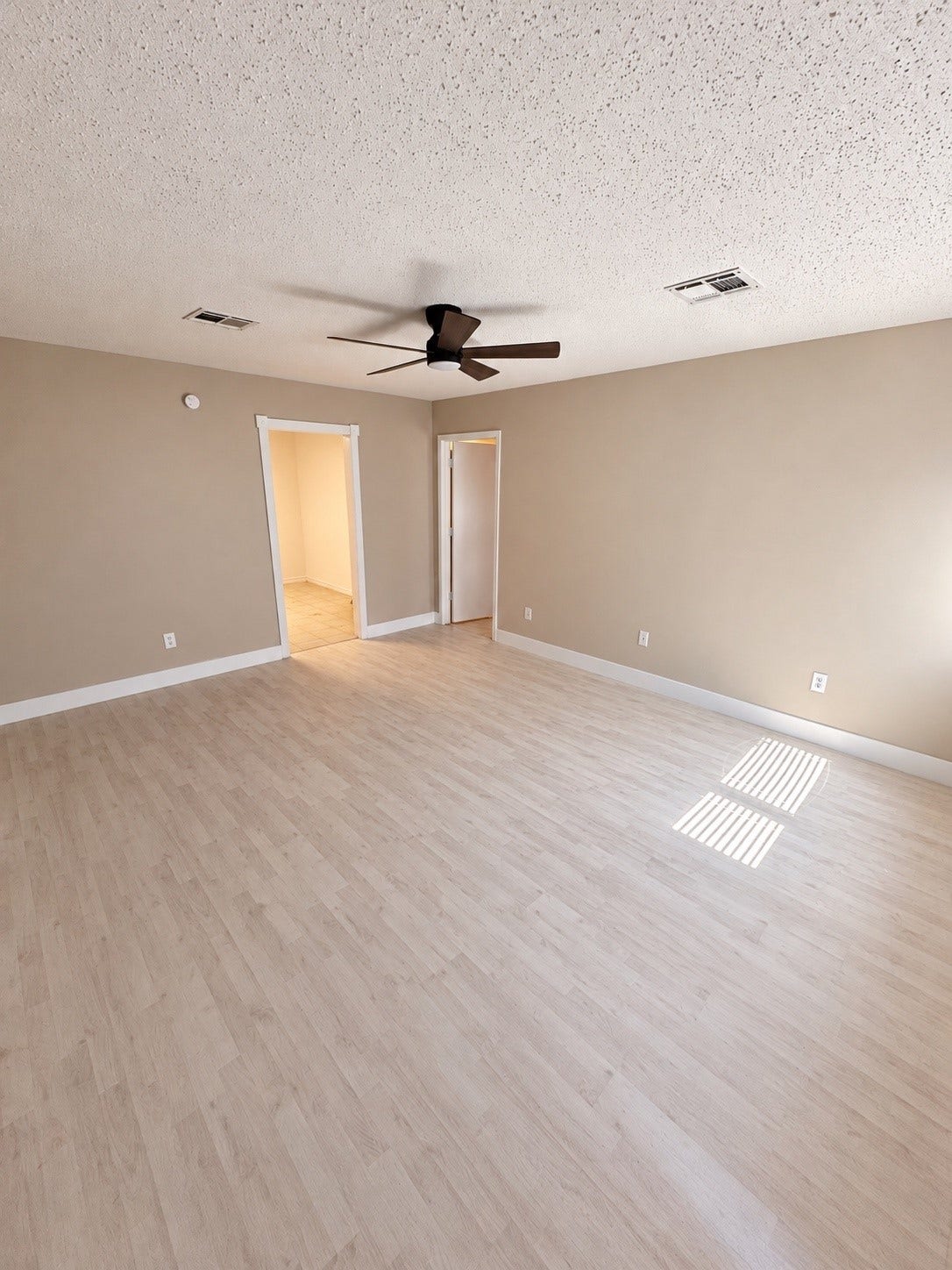Spacious room with light wood flooring and neutral walls, featuring a modern ceiling fan and ample natural light from a nearby window.