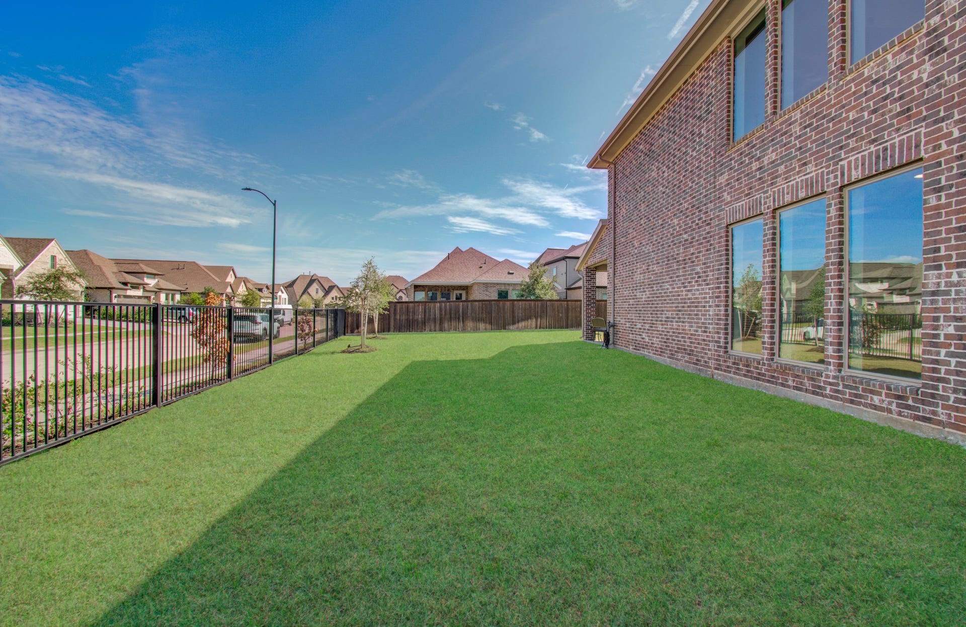 Spacious backyard with lush green lawn, bordered by a black metal fence, and large windows on a brick exterior offering ample natural light.