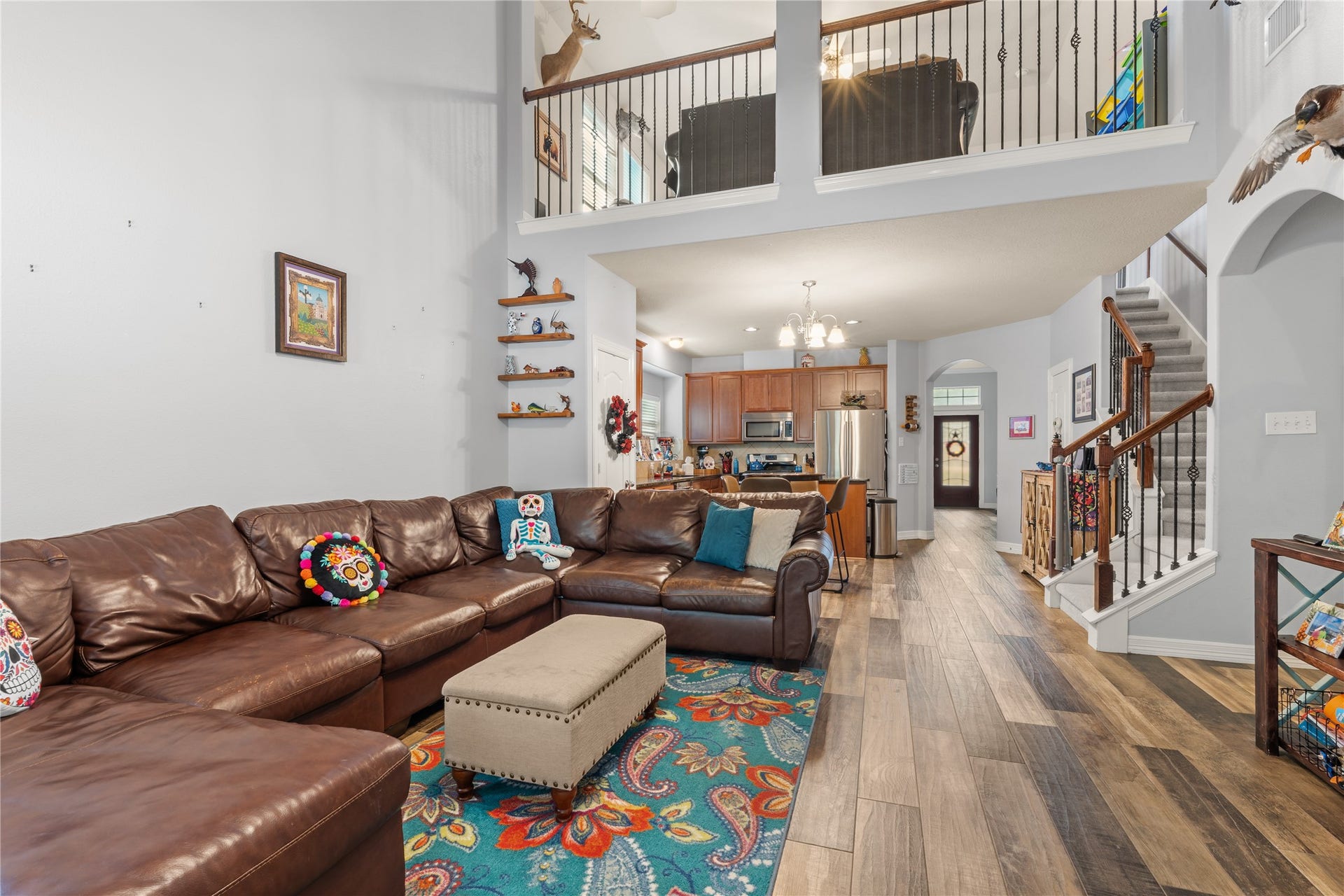 20184 Bitter Root Drive, Porter, TX 77365 Spacious living area with high ceilings, featuring a cozy leather sectional and colorful decor. The open-concept design flows seamlessly into the kitchen, boasting modern appliances and warm wooden cabinetry. An inviting staircase leads to an upper loft, enhancing the home's airy, welcoming feel.