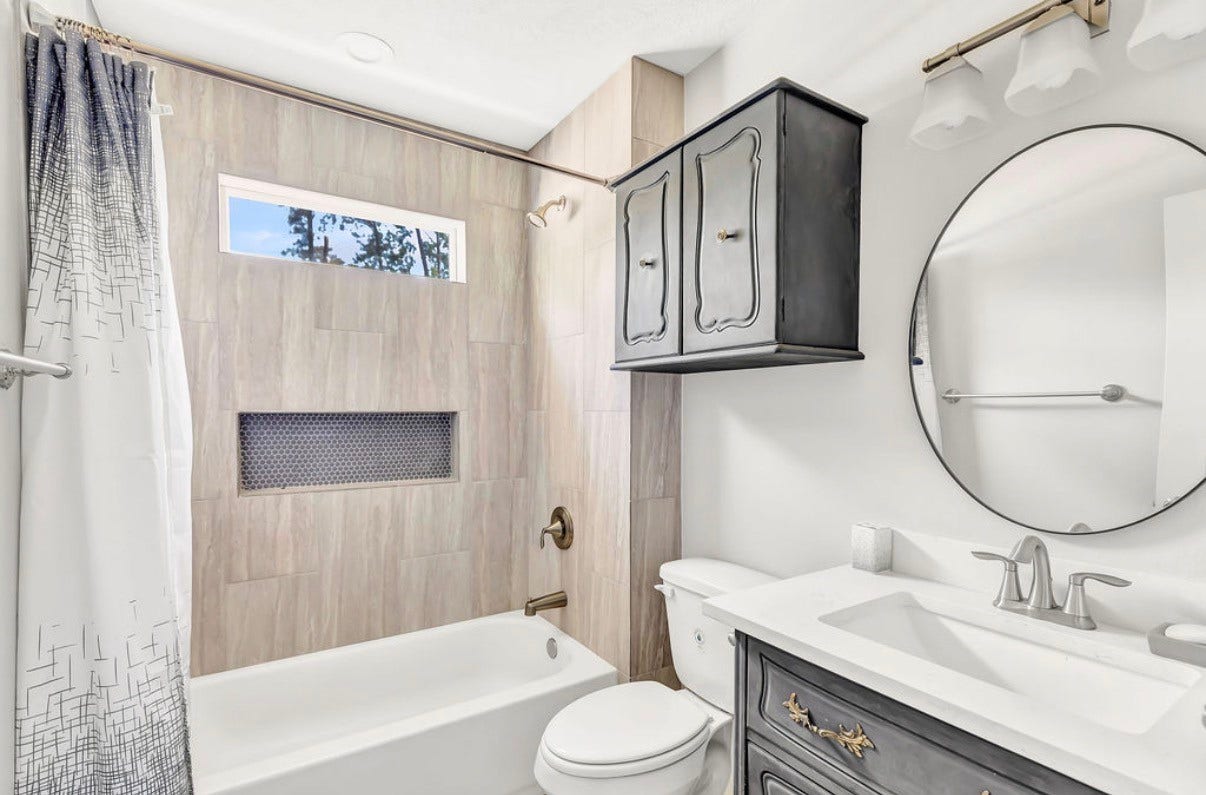 Experience modern elegance in this chic bathroom, featuring a sleek vanity with elegant fixtures, a stylish oval mirror, and a classic shower-tub combo framed by sophisticated tile work and a convenient built-in niche.