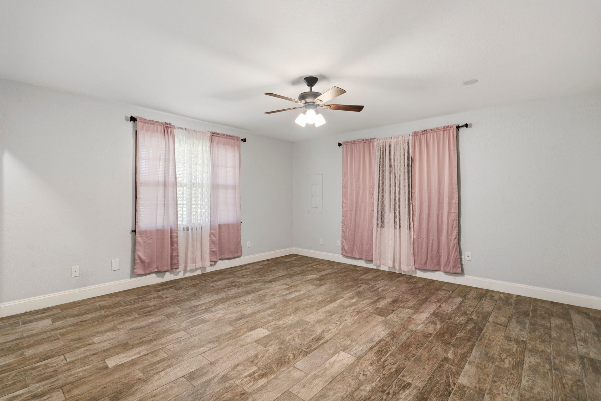 16101 Bluebird Street, Santa Fe, TX 77517 Spacious secondary room with warm wood-look flooring, soft gray walls, and ample natural light through large windows adorned with pink and white curtains. A ceiling fan adds a stylish touch.