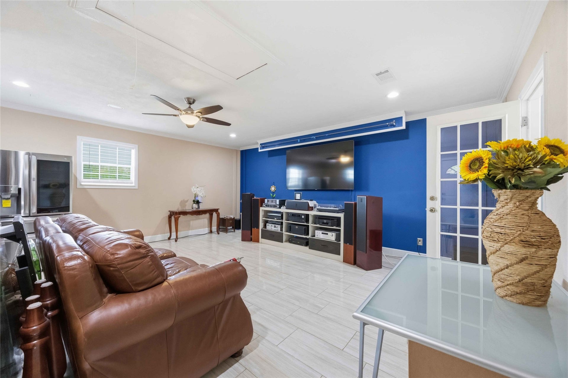 12075 Fm 359 Road, Hempstead, TX 77445 Spacious entertainment room/converted garage with sleek tiled flooring, a large wall-mounted TV, and modern media setup. Features a cozy leather couch, ceiling fan, and natural light from a window and glass door. Bright blue accent wall adds a pop of color.