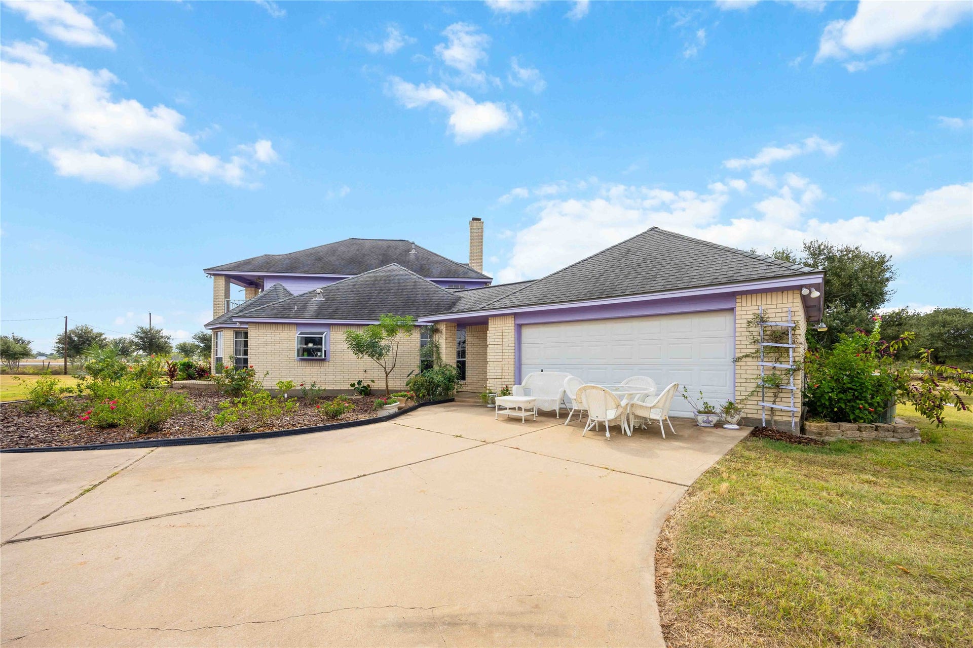 12075 Fm 359 Road, Hempstead, TX 77445 This is a charming two-story home with a spacious driveway leading to an attached garage. The exterior features light brick with purple accents, and the front yard is landscaped with plants. A patio area with seating is present, offering a welcoming outdoor space.