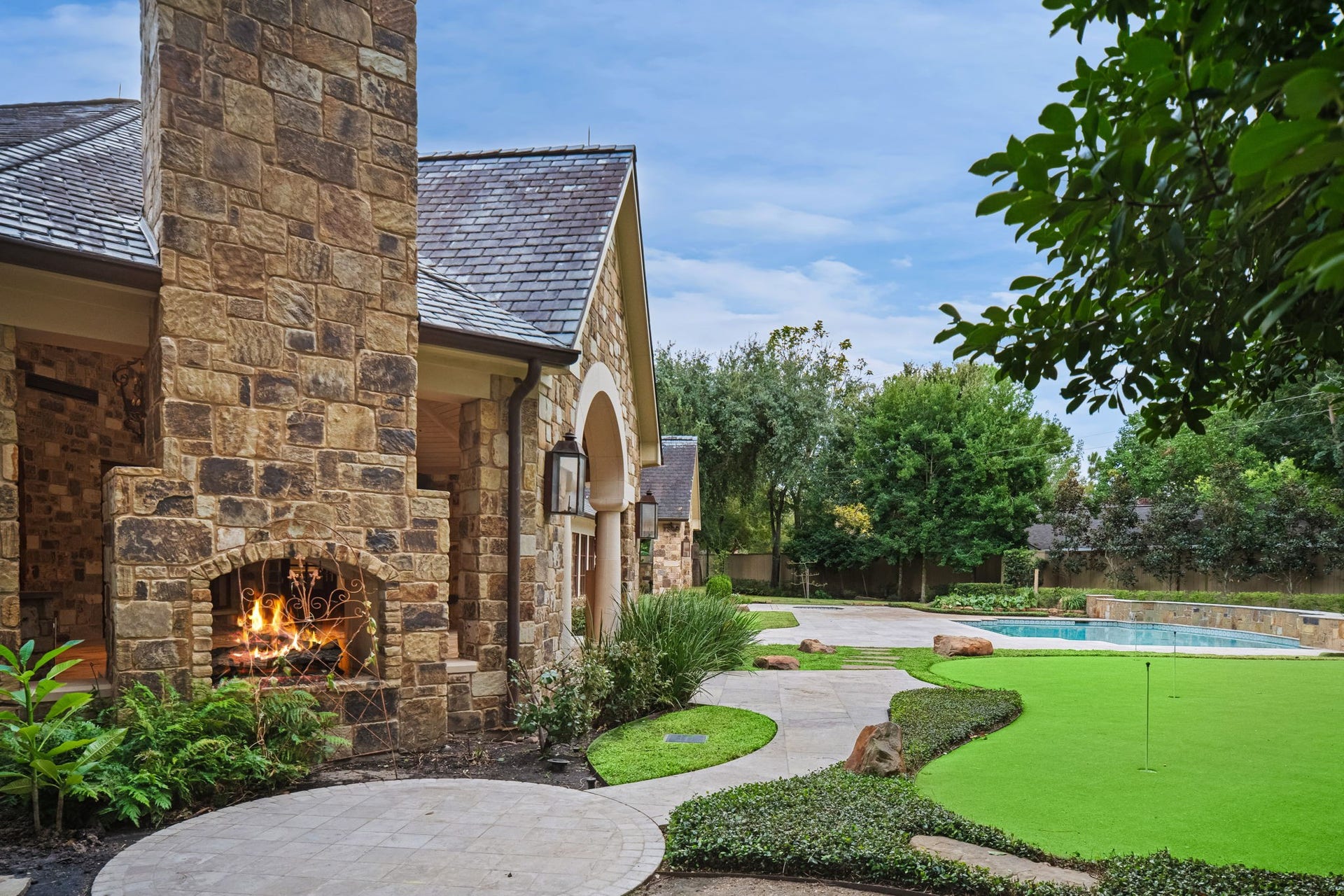 1 Gage Court, Houston, TX 77024 A closer view of the eight-hole putting green shows how the golf amenity is integrated directly into the landscaped backyard. The dual-sided fireplace at the outdoor pavilion and the homes stone architecture frame the green within the private grounds.