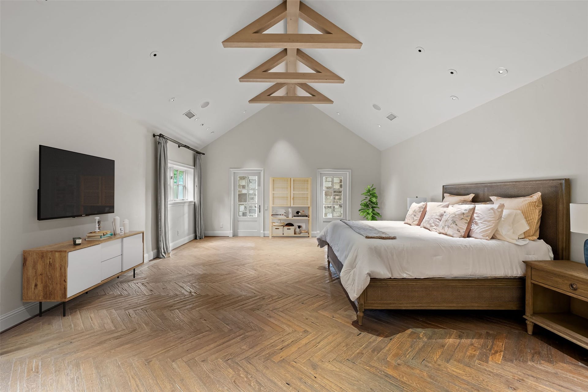 1 Gage Court, Houston, TX 77024 The primary bedroom features hand-scraped French white oak flooring laid in a herringbone pattern beneath a vaulted ceiling with exposed wood trusses. Generous proportions allow for a full bedroom suite with additional seating.