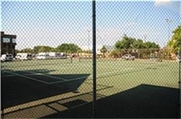 6001 Reims Road, #1202, Houston, TX 77036 The photo shows a fenced tennis court adjacent to a parking area, surrounded by trees and a building, ideal for recreational activities.