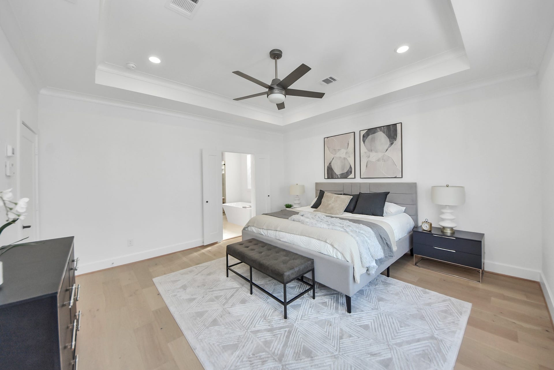 927 Fisher Street, #B, Houston, TX 77018 This spacious, modern bedroom invites relaxation with a neutral color palette, tray ceiling, recessed lighting, and a ceiling fan. A large bed with stylish bedding and matching nightstands anchors the room, while a contemporary rug adds warmth. The room connects seamlessly to an en-suite bathroom, enhancing the suites luxurious feel.