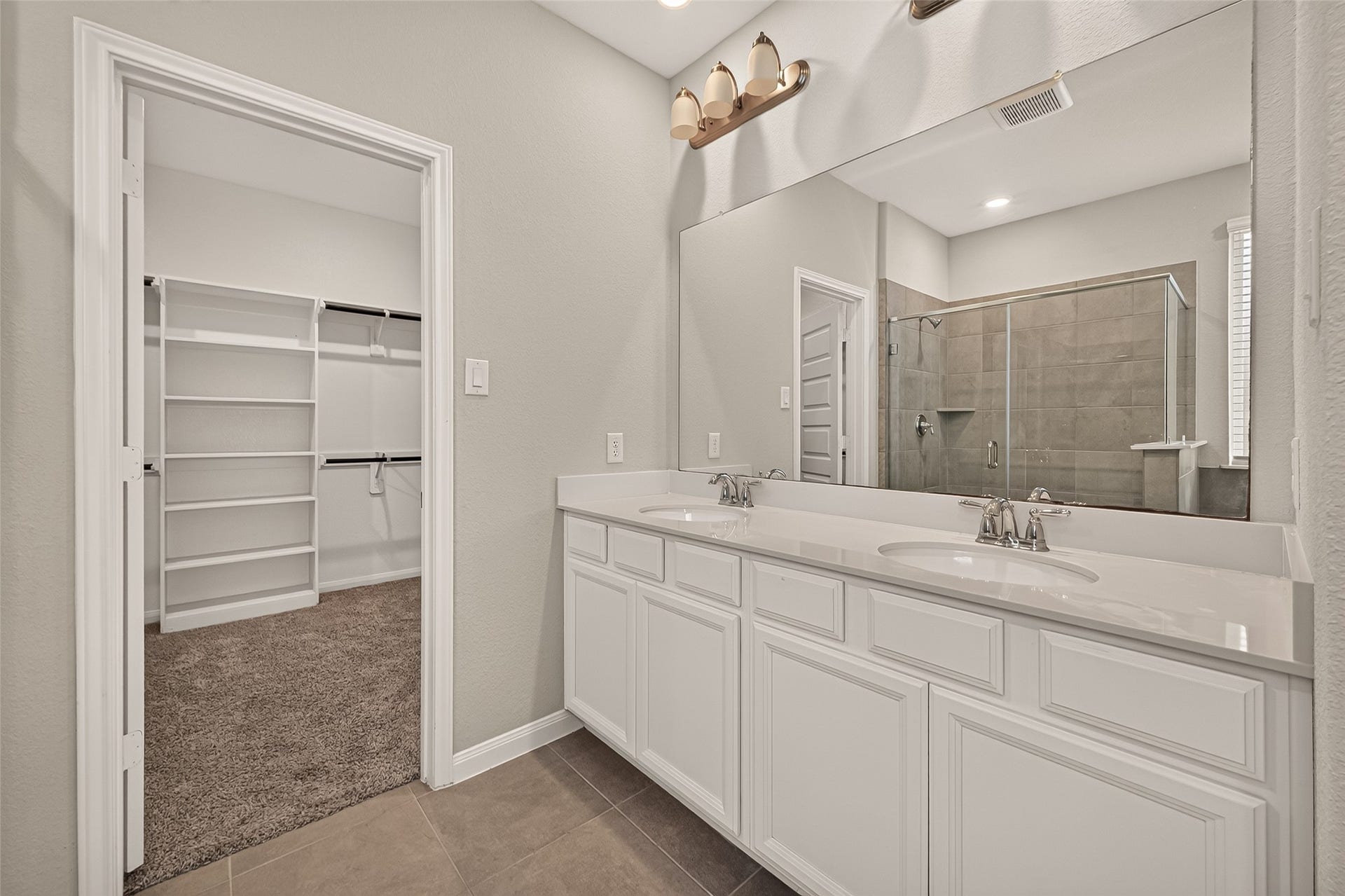 Elegant bathroom featuring a spacious double vanity, large mirror, and modern light fixtures, leading to a roomy walk-in closet and a sleek, glass-enclosed shower.
