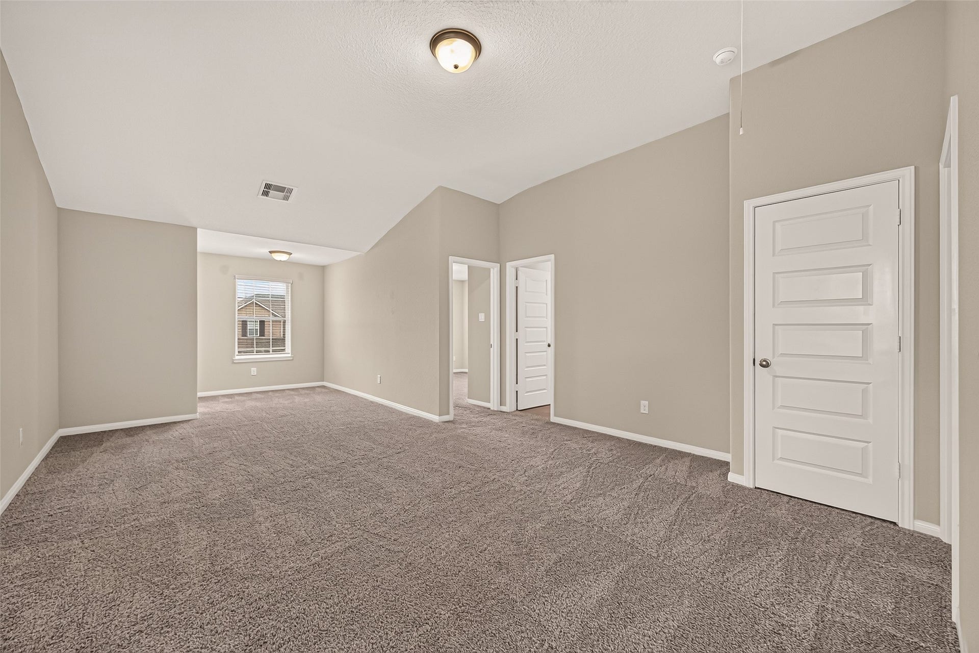 Spacious game room with plush carpeting, neutral walls, and ample natural light from a large window, complemented by clean, modern doors and a vaulted ceiling.