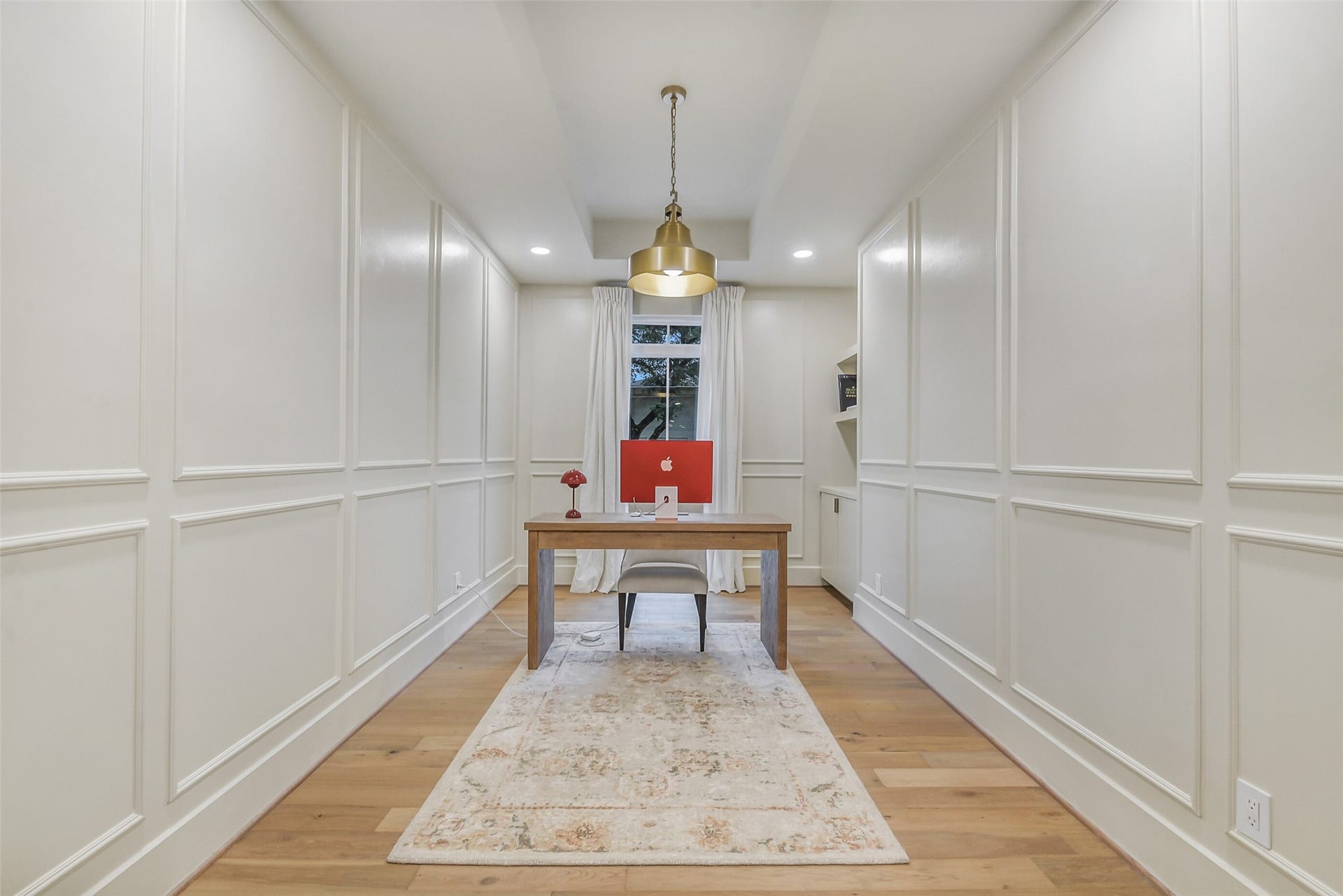 9411 Millbury Drive, Houston, TX 77096 Millwork paneling covers every wall in this refined home office, adding architectural detail and a tailored, sophisticated feel. A statement brass pendant light hangs above the desk, with a sunlit window and built-in shelving completing the space.