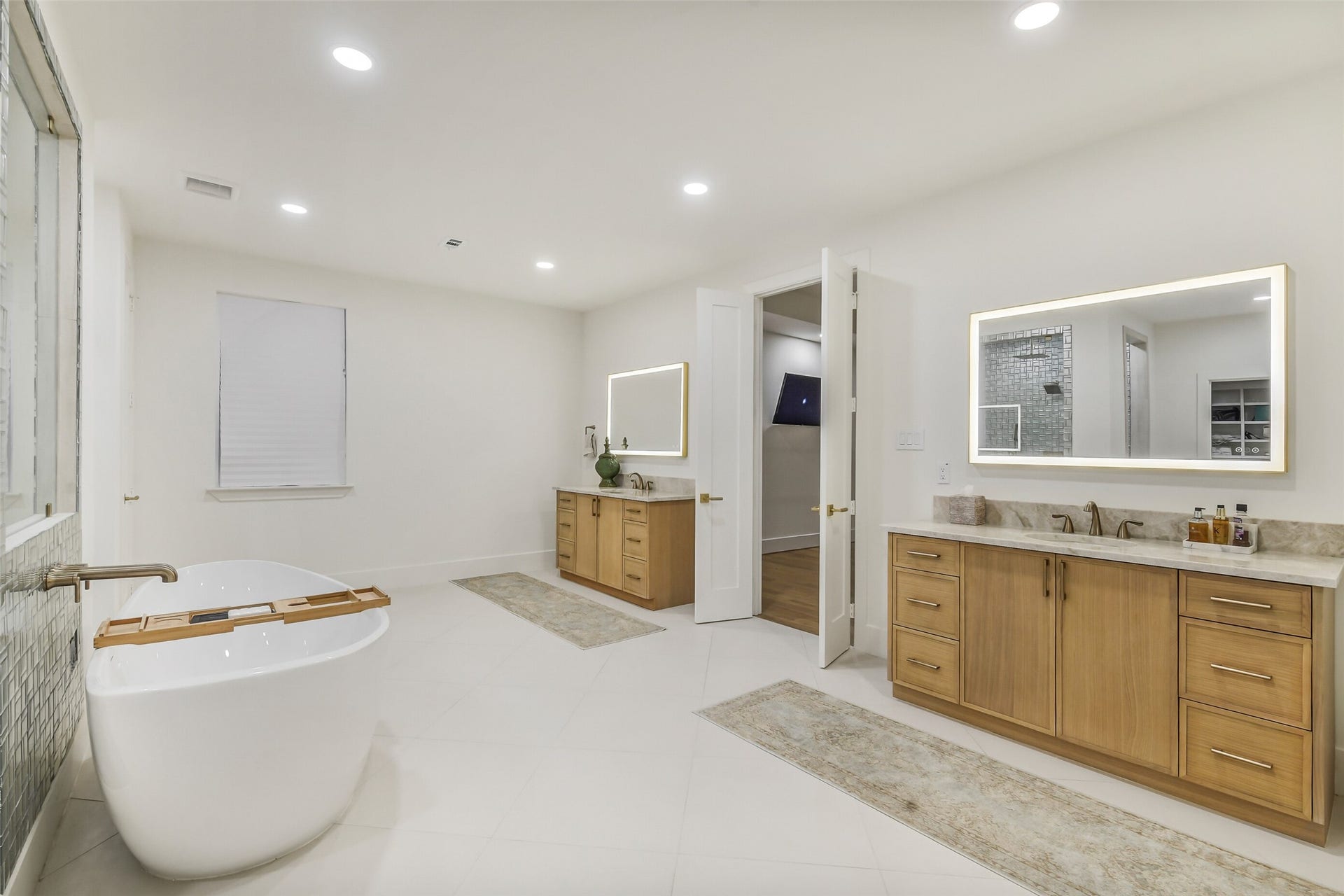 9411 Millbury Drive, Houston, TX 77096 A spa-like primary bath featuring a freestanding soaking tub, two separate vanities with backlit mirrors, and a walk-in tiled shower. Warm wood cabinetry, quartz countertops, and thoughtful details throughout create a luxurious, private retreat.