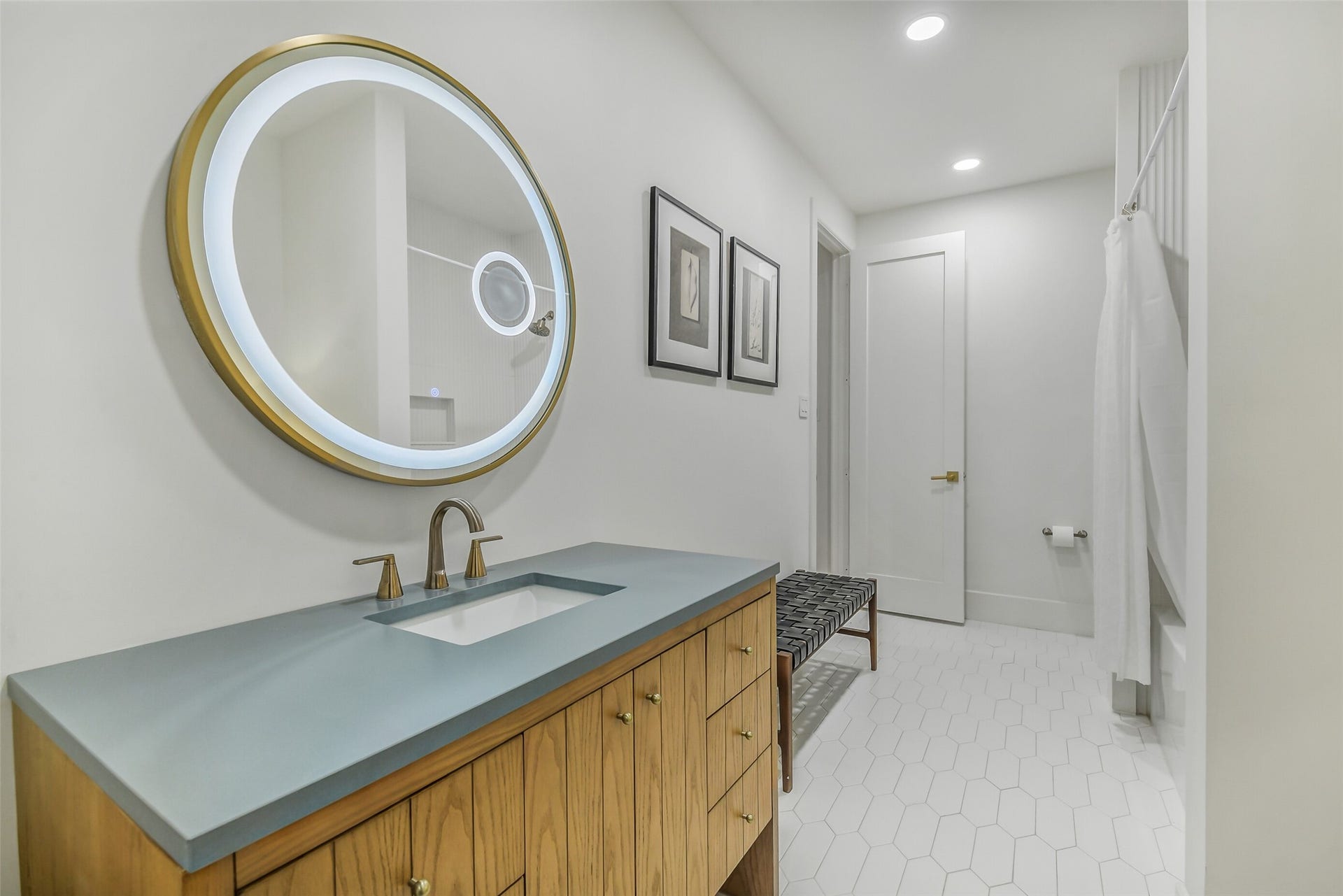 9411 Millbury Drive, Houston, TX 77096 Gorgeous bath featuring a striking round backlit mirror in a brass frame, a blue concrete-look countertop, and warm wood cabinetry with brass hardware. Hexagon tile flooring and a woven bench add character throughout.