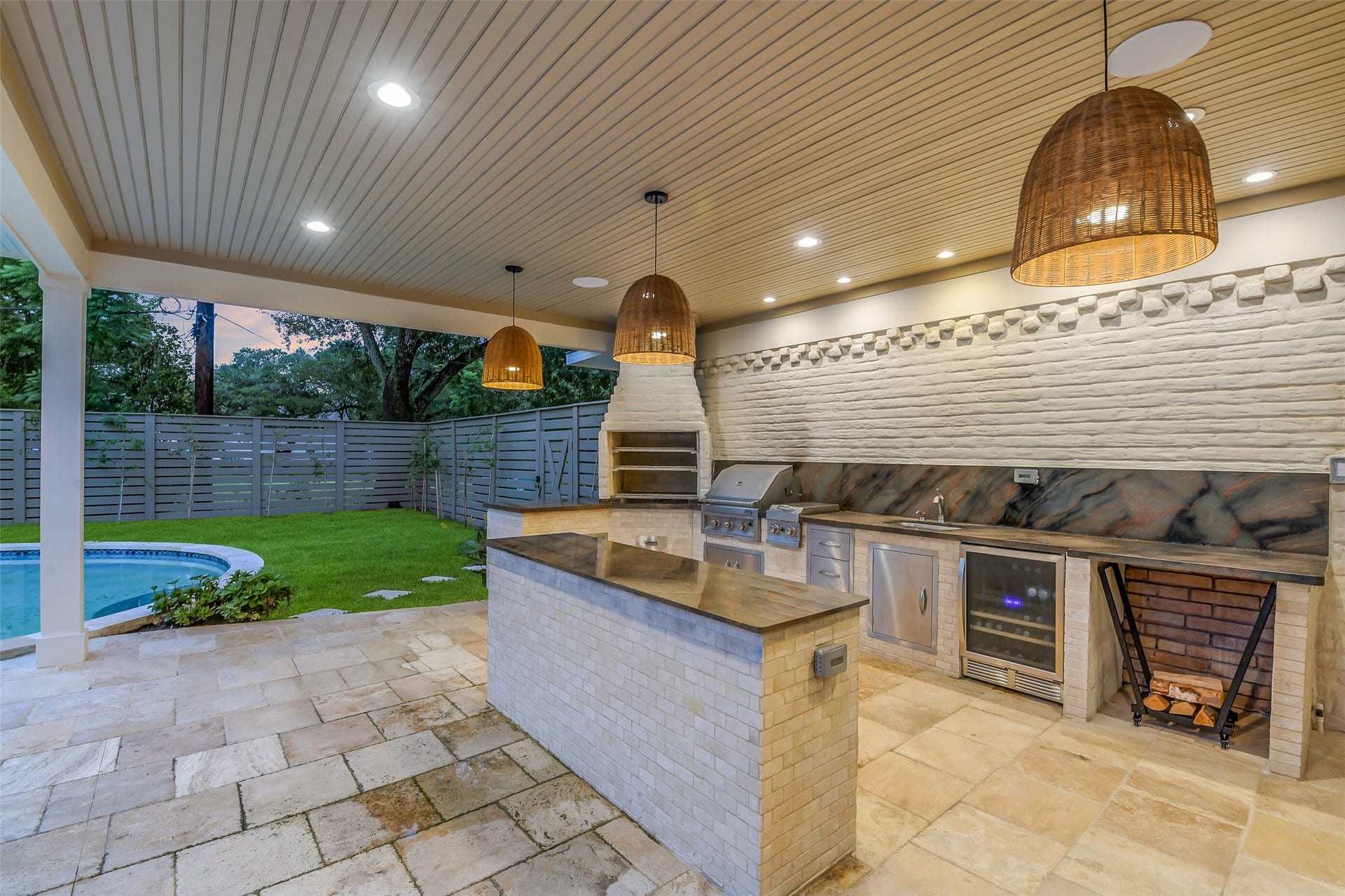 9411 Millbury Drive, Houston, TX 77096 A true outdoor entertainer's dream, this fully equipped kitchen features a built-in grill, wine fridge, sink, and generous prep counters, all set beneath a woven pendant-lit wood ceiling. A brick pizza oven and wood-storage nook add rustic charm, while the space overlooks the pool and privately fenced backyard.