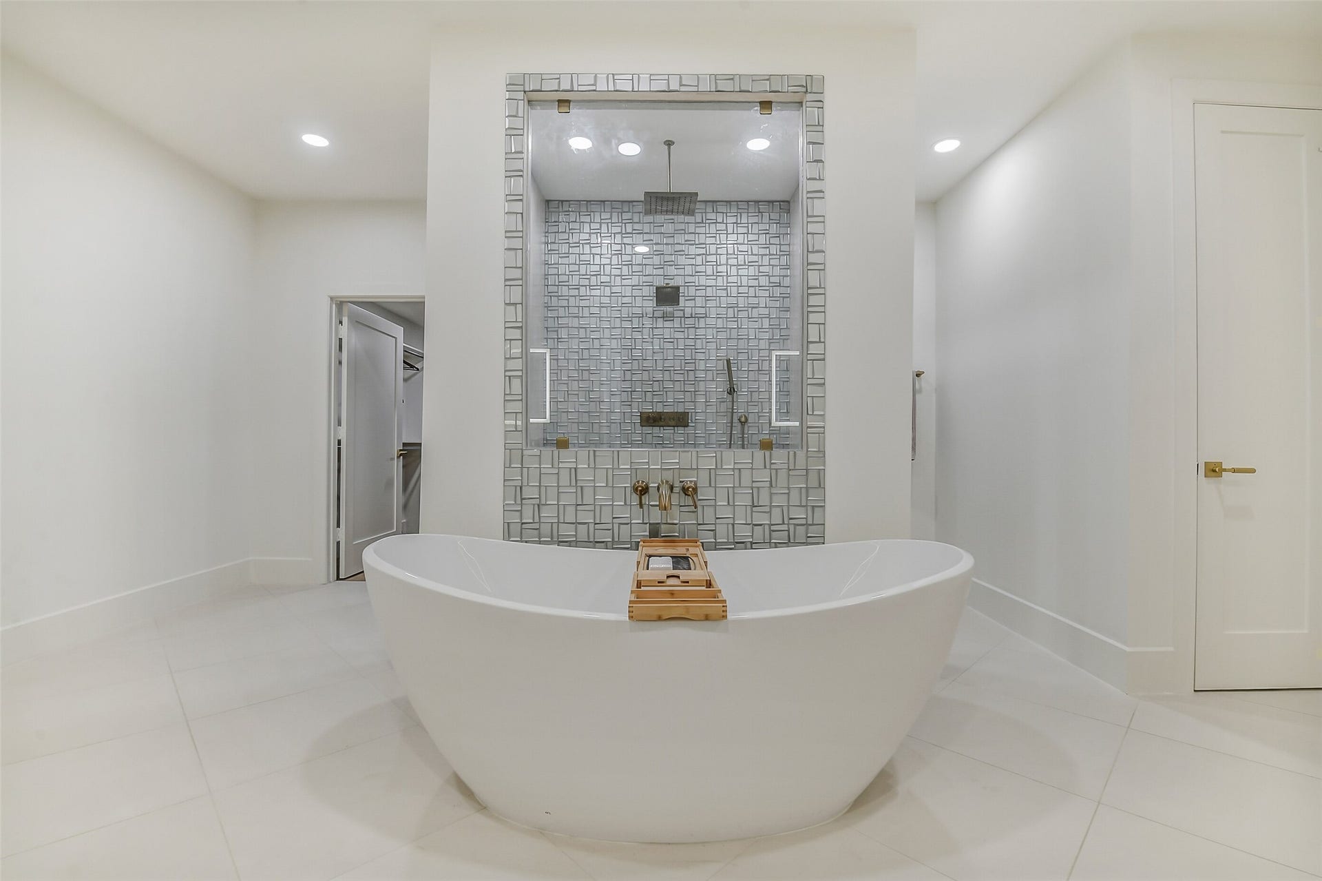 9411 Millbury Drive, Houston, TX 77096 An oversized soaking tub sits front and center, framed by a stunning textured tile shower with a rain showerhead and handheld sprayer. Sleek brass fixtures and a spa-like walk-in closet nearby complete this serene, luxurious retreat.
