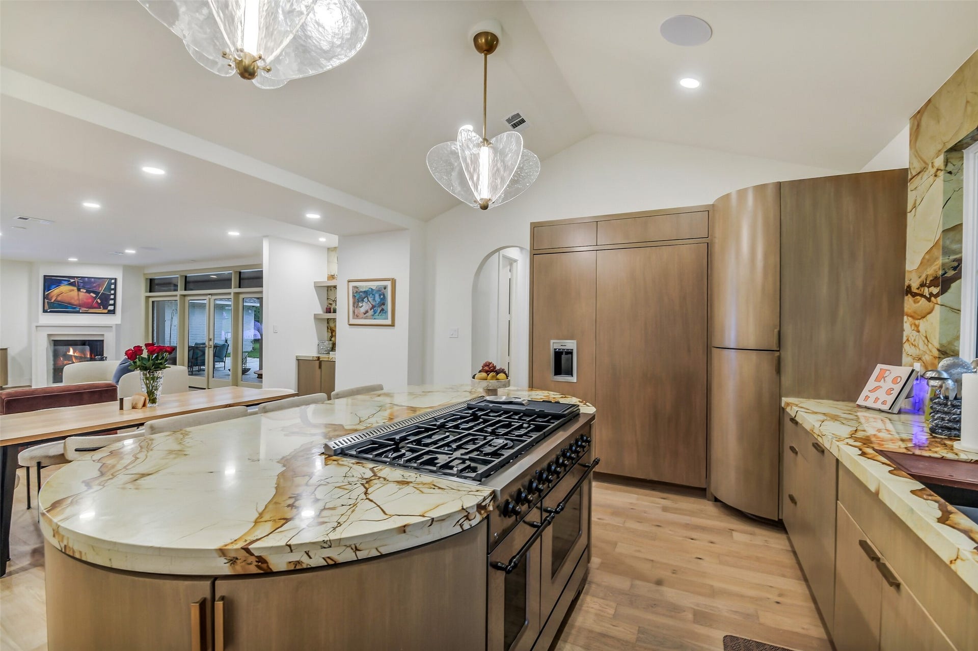 9411 Millbury Drive, Houston, TX 77096 A show-stopping curved island anchors this chef's kitchen, wrapped in dramatic natural stone and fitted with a professional-grade gas range. Custom wood cabinetry conceals the built-in refrigerator, while sculptural glass pendants and a vaulted ceiling add elegance overhead.