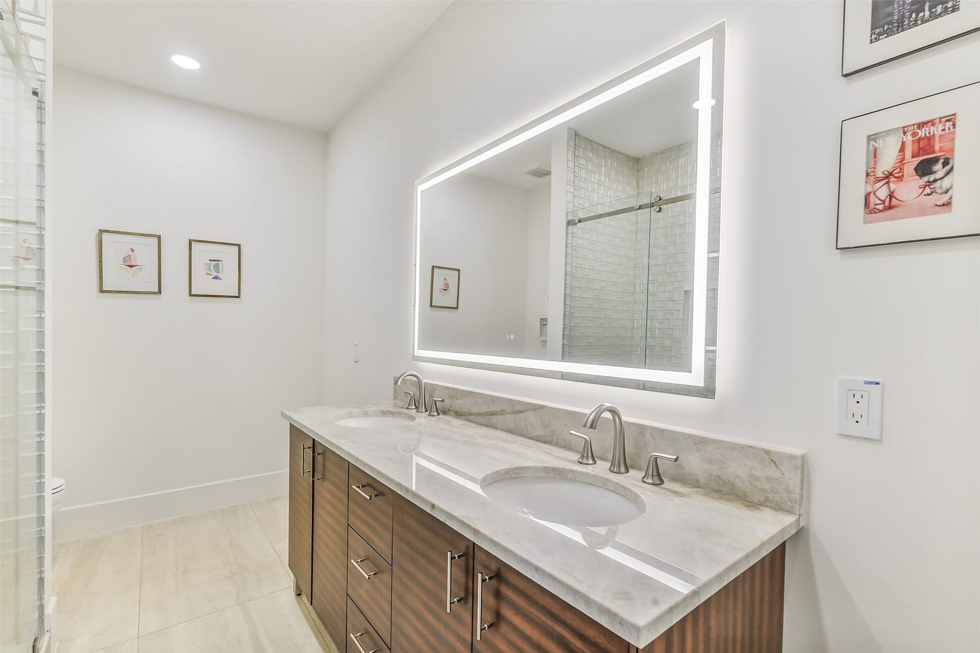 9411 Millbury Drive, Houston, TX 77096 A sleek en-suite featuring a dual-sink vanity with rich wood cabinetry, a backlit LED mirror, and a glass-enclosed tiled shower. Stone countertops and modern fixtures give this bath a polished, upscale feel.