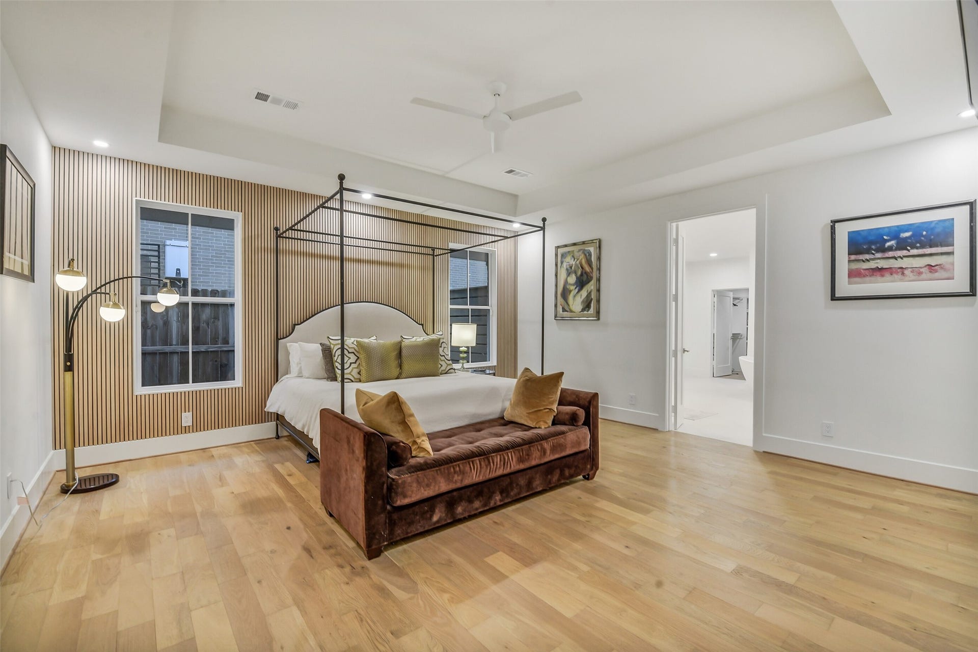 9411 Millbury Drive, Houston, TX 77096 A spacious primary suite featuring a slatted wood accent wall behind the bed and a tray ceiling that adds architectural depth. A sleek canopy bed frame, plush velvet bench, and large windows create a warm, sophisticated retreat, with direct access to the en-suite bath just beyond.
