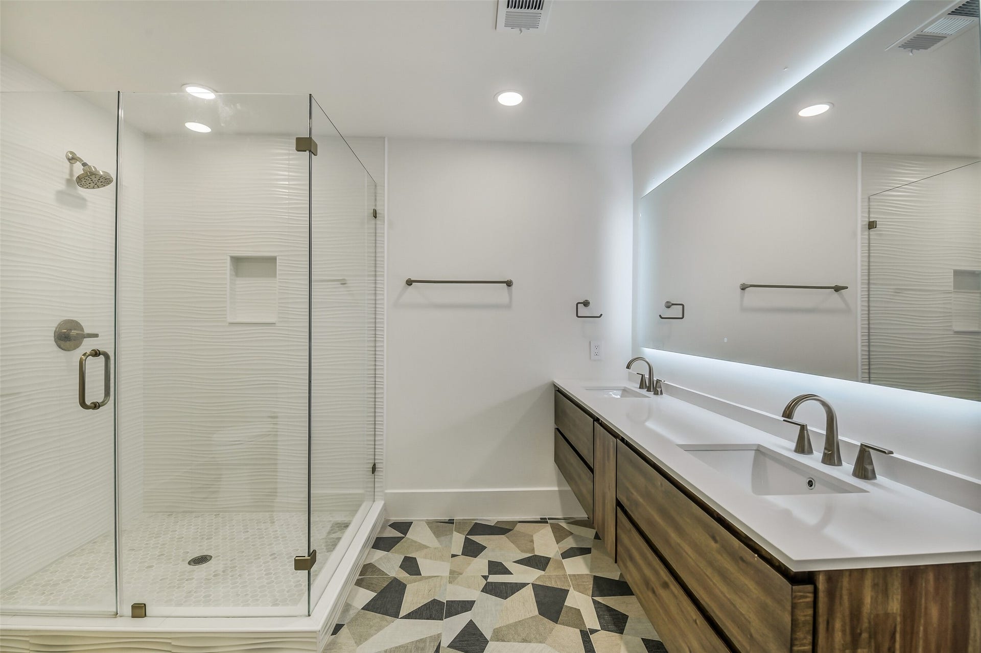9411 Millbury Drive, Houston, TX 77096 A modern bathroom featuring a glass-enclosed shower with textured tile, a floating dual-sink vanity, and a striking geometric tile floor. A backlit mirror and warm wood cabinetry add a sleek, contemporary finish.