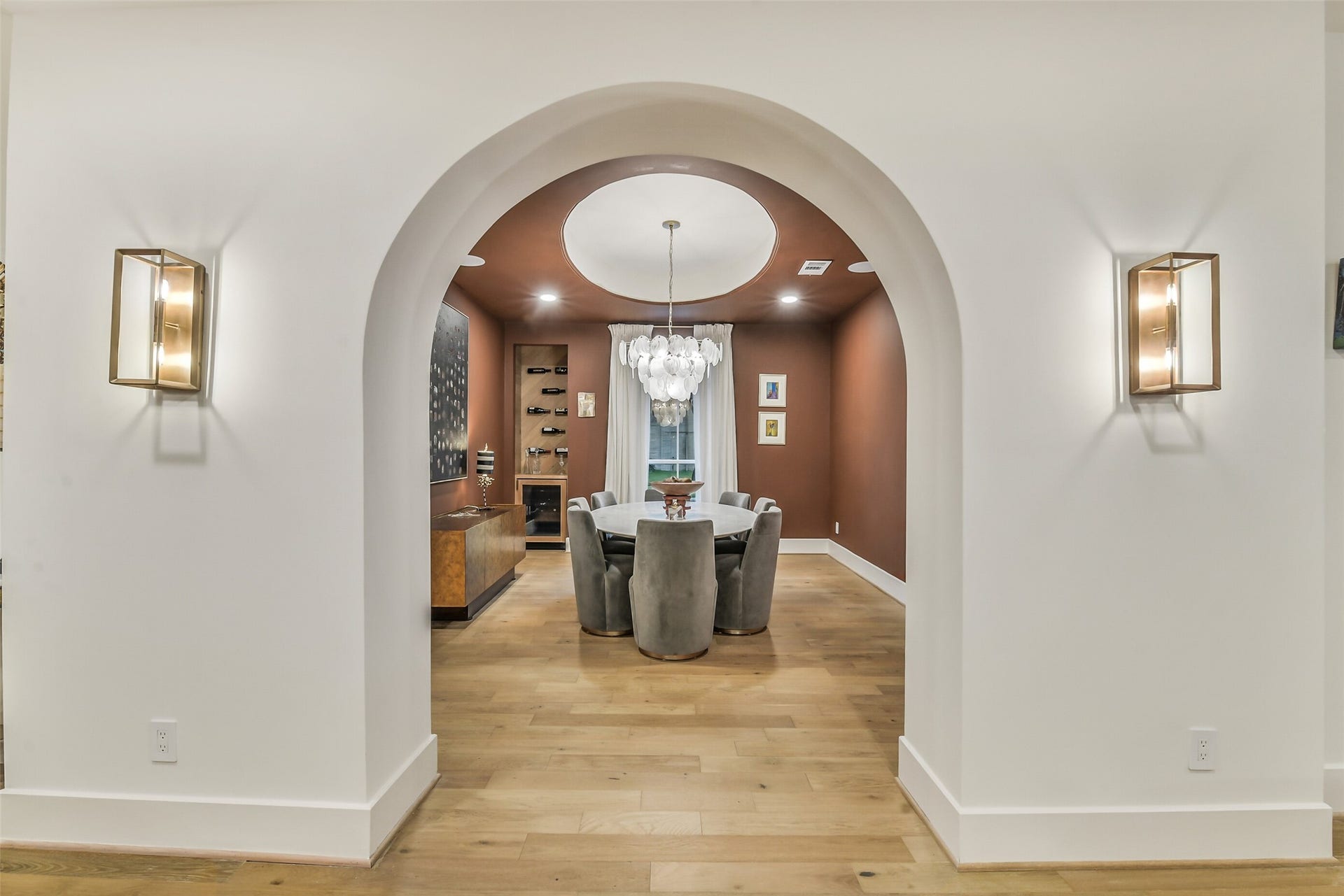 9411 Millbury Drive, Houston, TX 77096 A bold arched entry framed by brass sconces leads into this moody, richly-hued dining room. A statement bubble chandelier hangs from a domed ceiling detail above the round table, while a built-in wine display and custom bar cabinet set the scene for elegant entertaining.