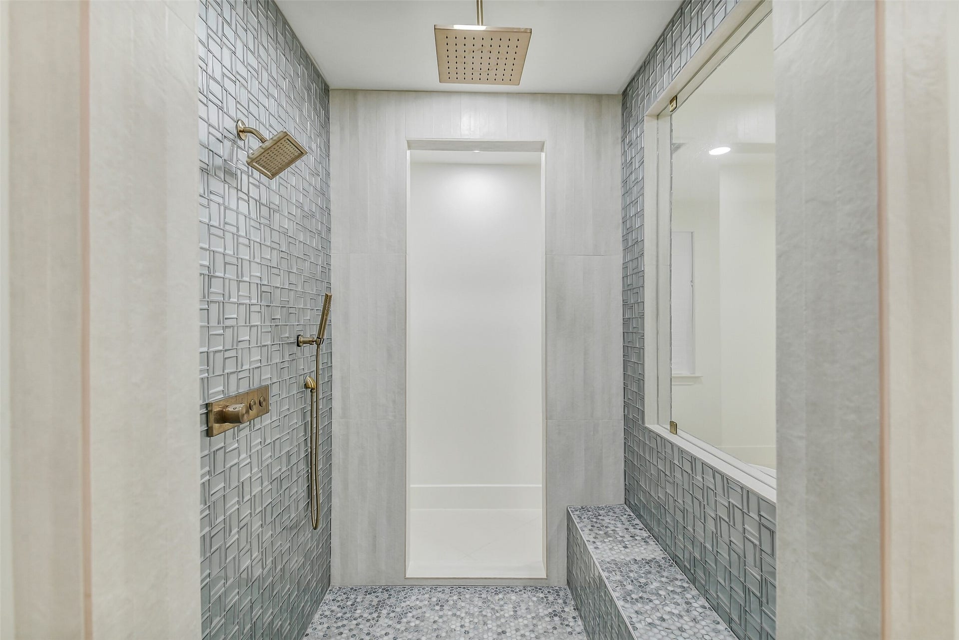 9411 Millbury Drive, Houston, TX 77096 An expansive walk-in shower featuring textured mosaic tile, a built-in bench, and dual showerheads a rain head overhead plus a handheld sprayer. Brushed brass fixtures and a pebble tile floor add a spa-inspired finishing touch.