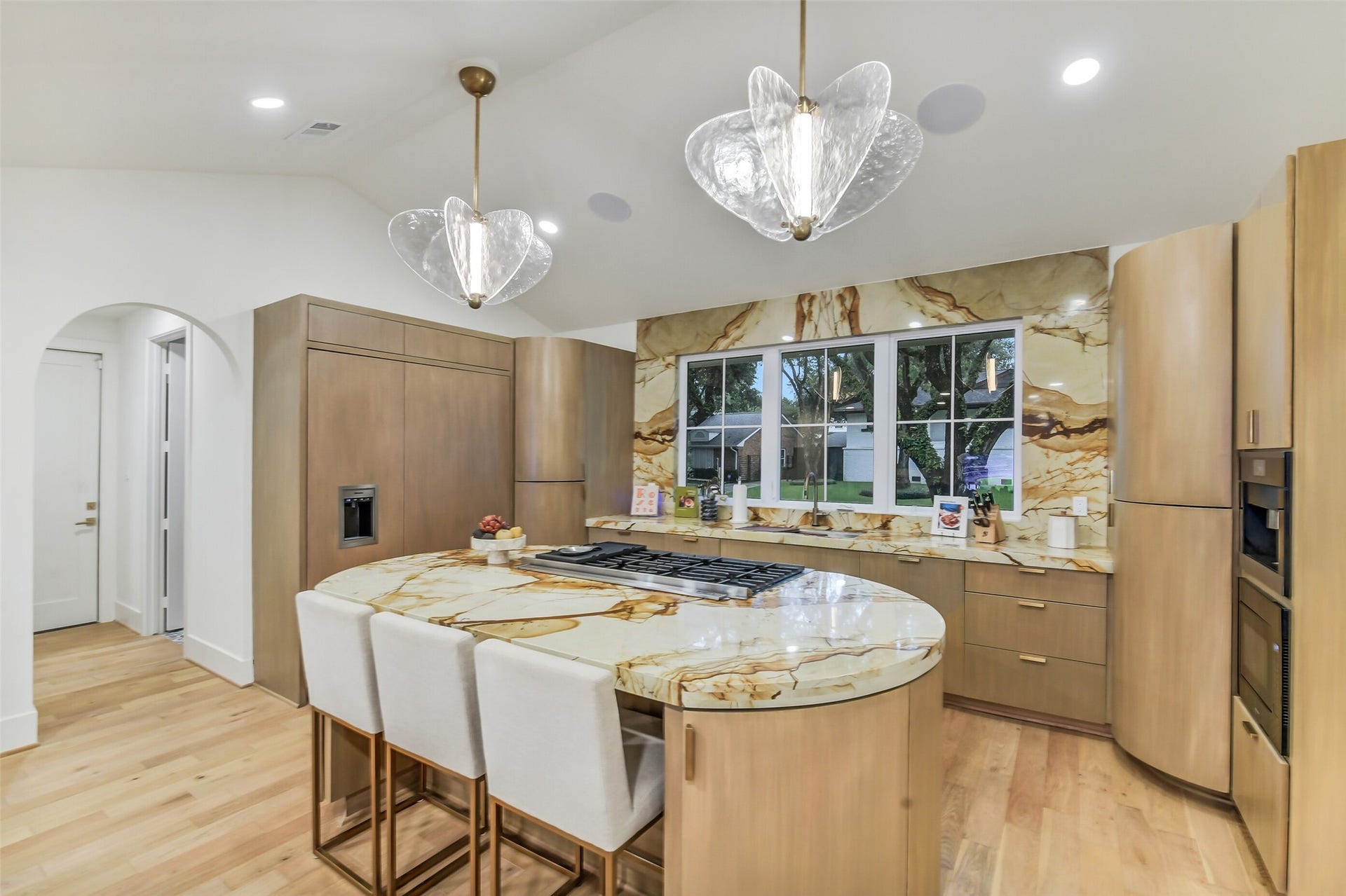 9411 Millbury Drive, Houston, TX 77096 A striking kitchen featuring dramatic stone-slab backsplash, warm wood cabinetry, and an oversized island with bar seating beneath sculptural glass pendant lighting. Paneled-front appliances and a sunny window over the sink complete this elegant, functional space.