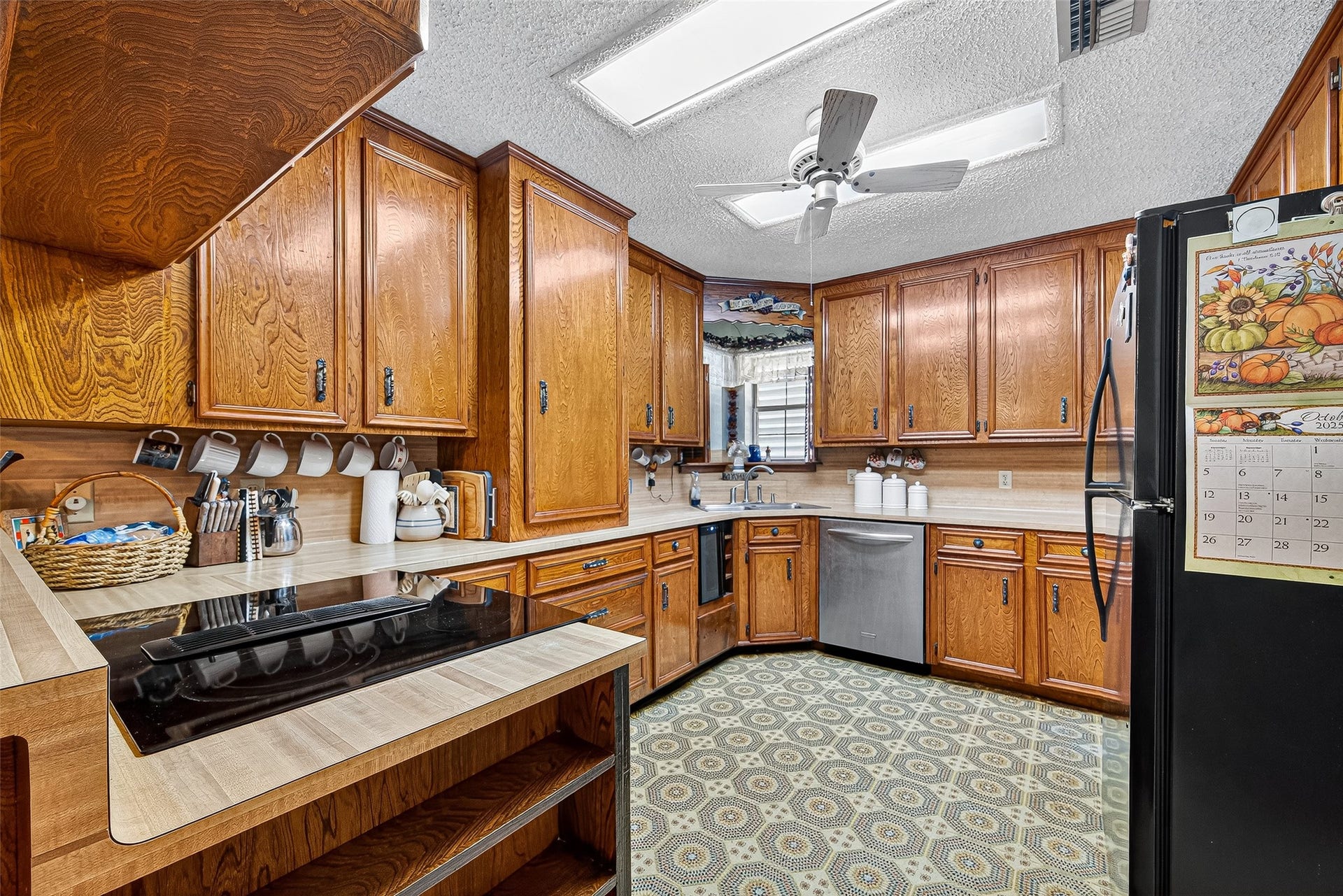 12021 7th 1/2 Street, Santa Fe, TX 77510 This kitchen features classic wooden cabinetry with ample storage, a modern cooktop, and a stainless steel dishwasher. It has a cozy atmosphere with patterned flooring, ceiling fan, and natural light from a window above the sink.