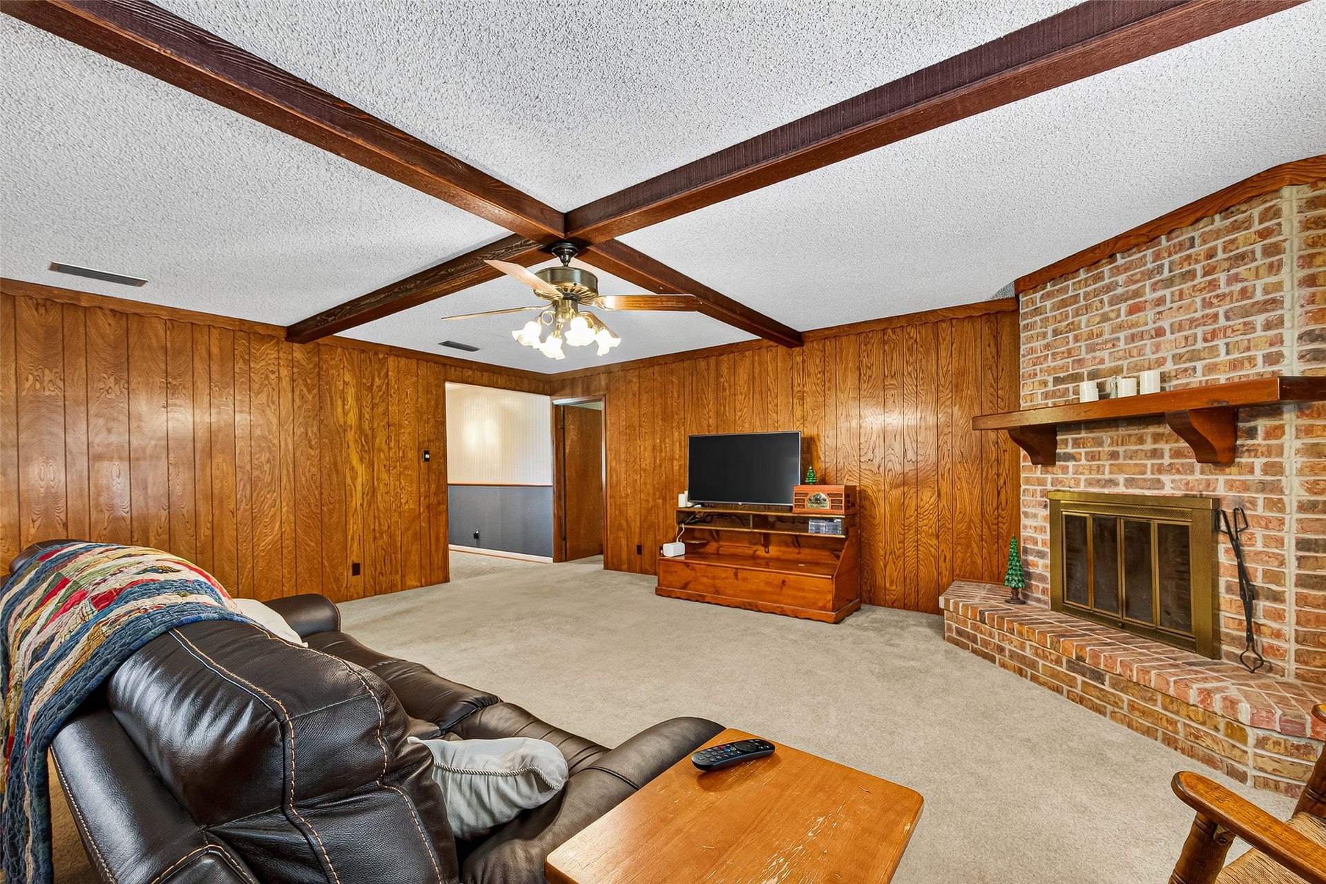 12021 7th 1/2 Street, Santa Fe, TX 77510 This cozy living room features wood-paneled walls and a charming brick fireplace. It has a ceiling fan, exposed beams, and ample space for seating, making it perfect for relaxation and gatherings.