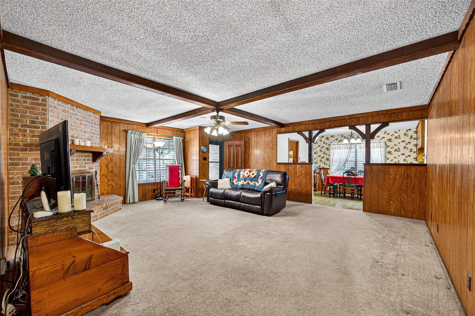 12021 7th 1/2 Street, Santa Fe, TX 77510 This cozy living room features wood-paneled walls, a brick fireplace, and beamed ceiling accents. It opens to a dining area with a large window, offering a warm and inviting space for gatherings.