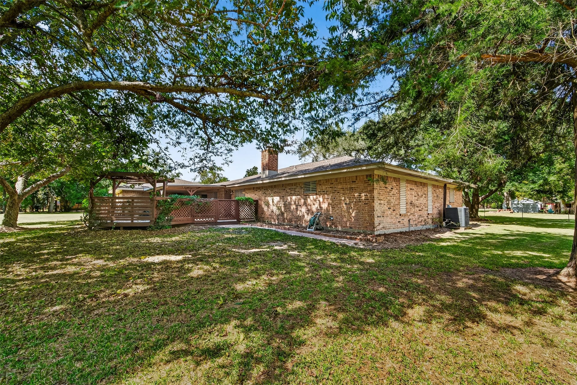 12021 7th 1/2 Street, Santa Fe, TX 77510 This photo shows the backyard of a brick house with a spacious lawn. There's a wooden deck or pergola attached to the house, offering a cozy outdoor space. Hot tub ready for installation. The setting is peaceful, with plenty of greenery.