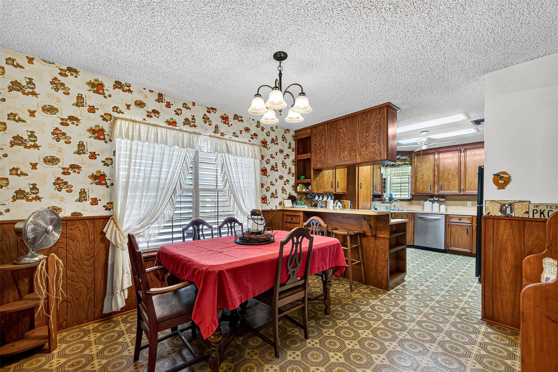 12021 7th 1/2 Street, Santa Fe, TX 77510 This cozy dining area features vintage floral wallpaper and wood paneling, creating a warm, nostalgic atmosphere. It has a spacious table, abundant natural light from large windows, and an open view into a well-equipped kitchen with wood cabinetry.