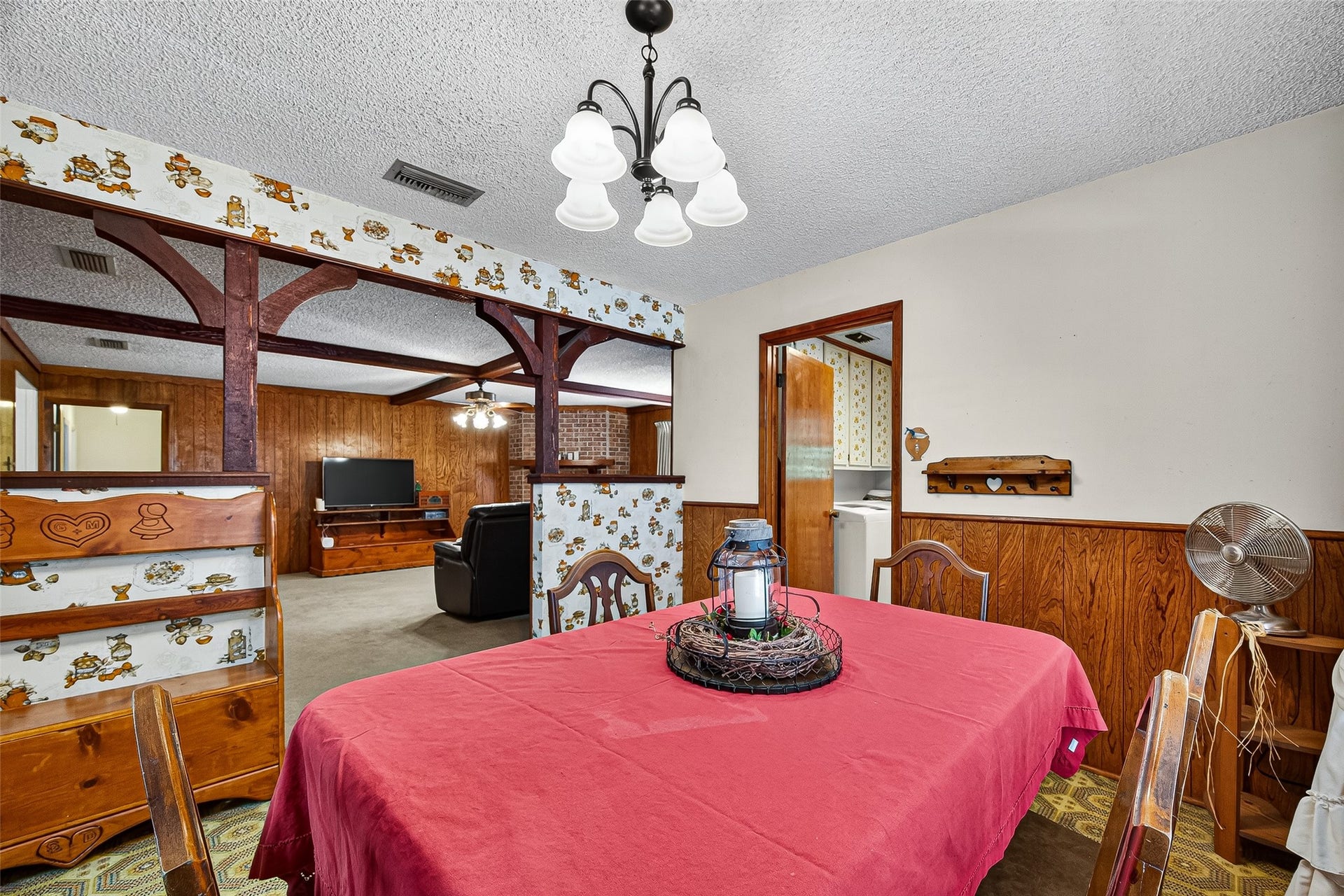 12021 7th 1/2 Street, Santa Fe, TX 77510 Cozy dining area with vintage charm, featuring wood paneling, patterned wallpaper, and decorative beams. The space is open to a living room with a TV, offering a warm and inviting atmosphere.