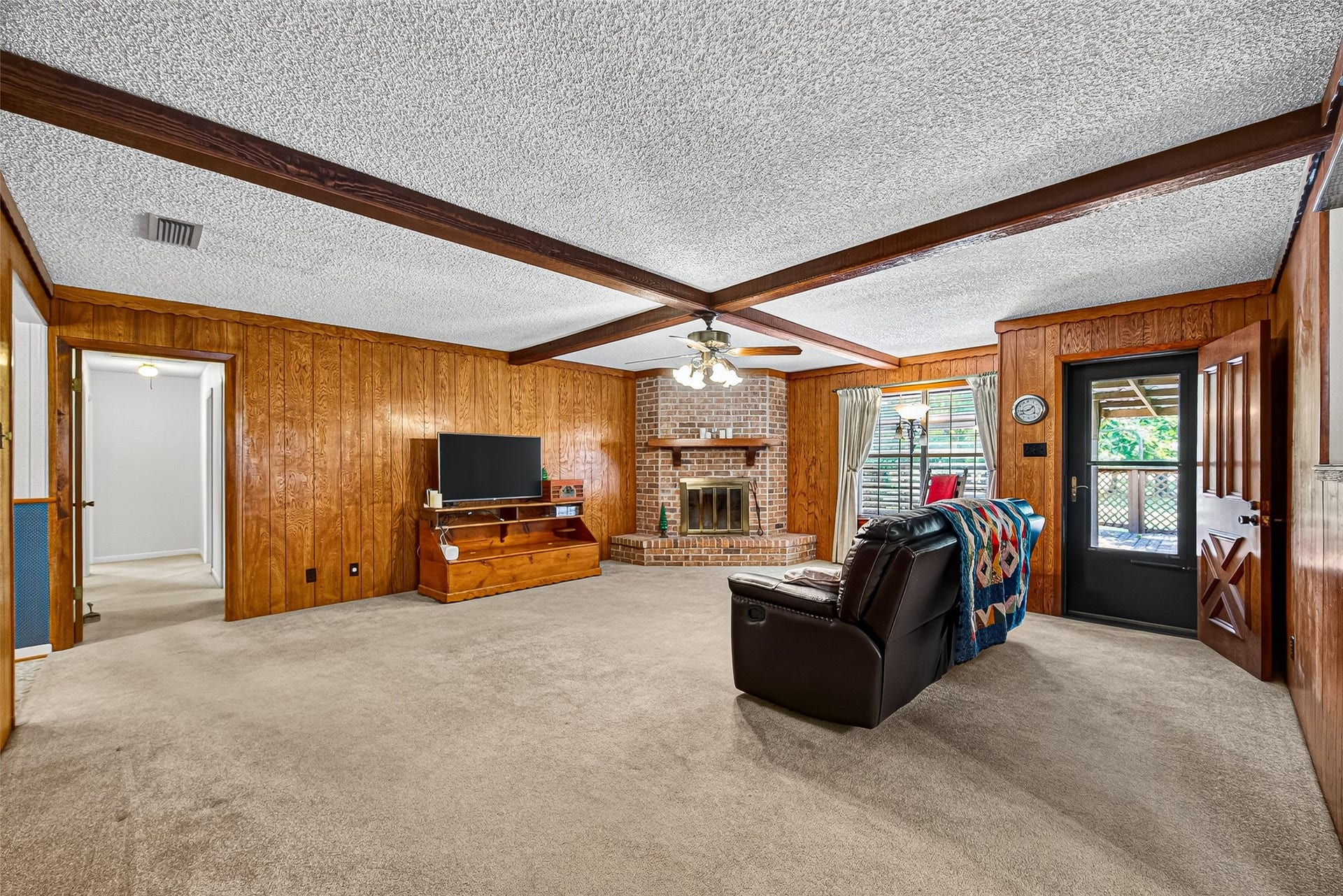 12021 7th 1/2 Street, Santa Fe, TX 77510 This cozy living room features wood-paneled walls, a brick fireplace, and a ceiling with exposed beams. It has carpeted flooring, a ceiling fan, and large windows allowing natural light. A door leads to an outdoor space.