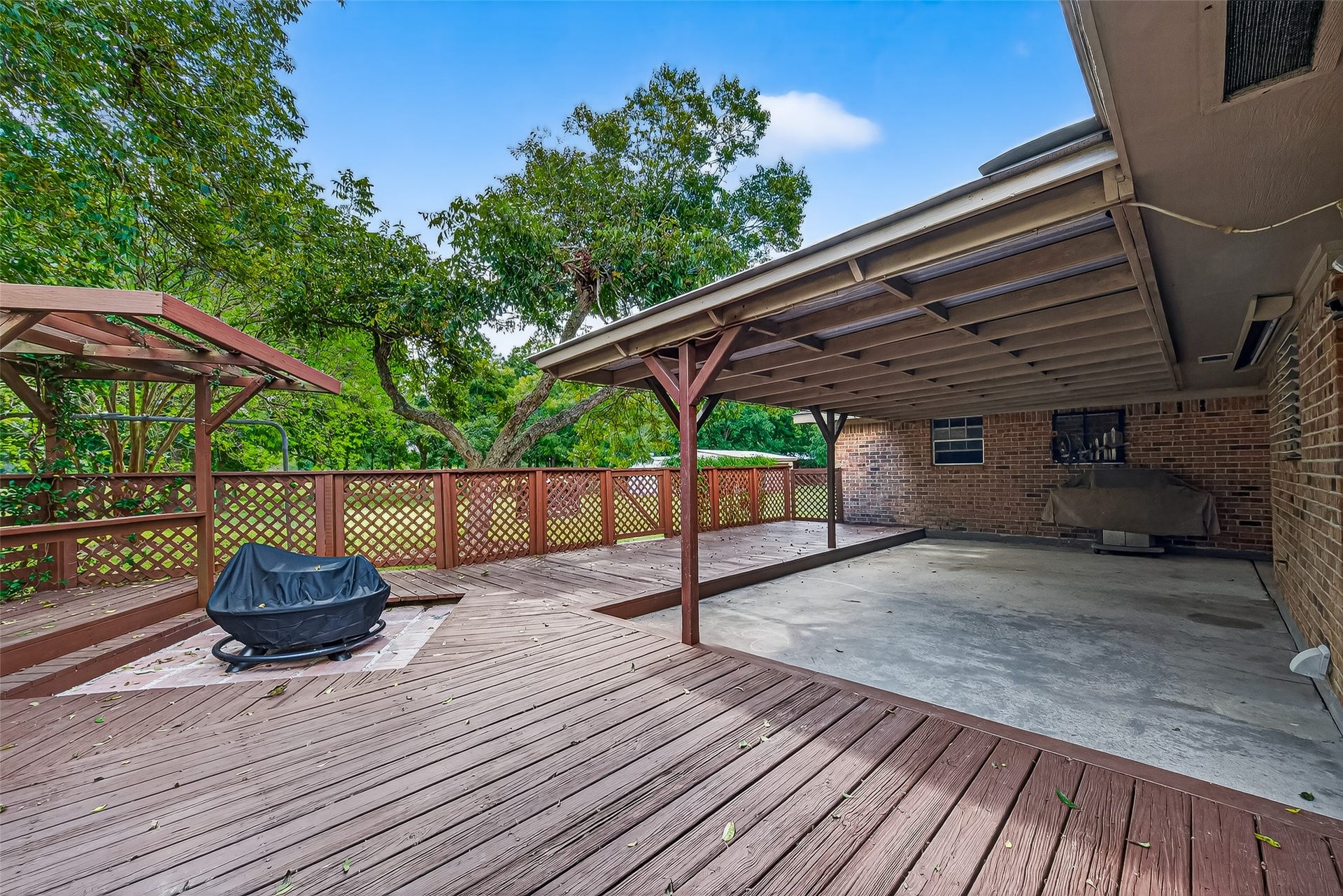 12021 7th 1/2 Street, Santa Fe, TX 77510 This photo shows a spacious backyard patio featuring a large wooden deck with a fire pit and a covered area for shade and ready for hot tub to be installed. The space is surrounded by lush greenery, providing a private and serene outdoor setting. Perfect for entertaining or relaxing.