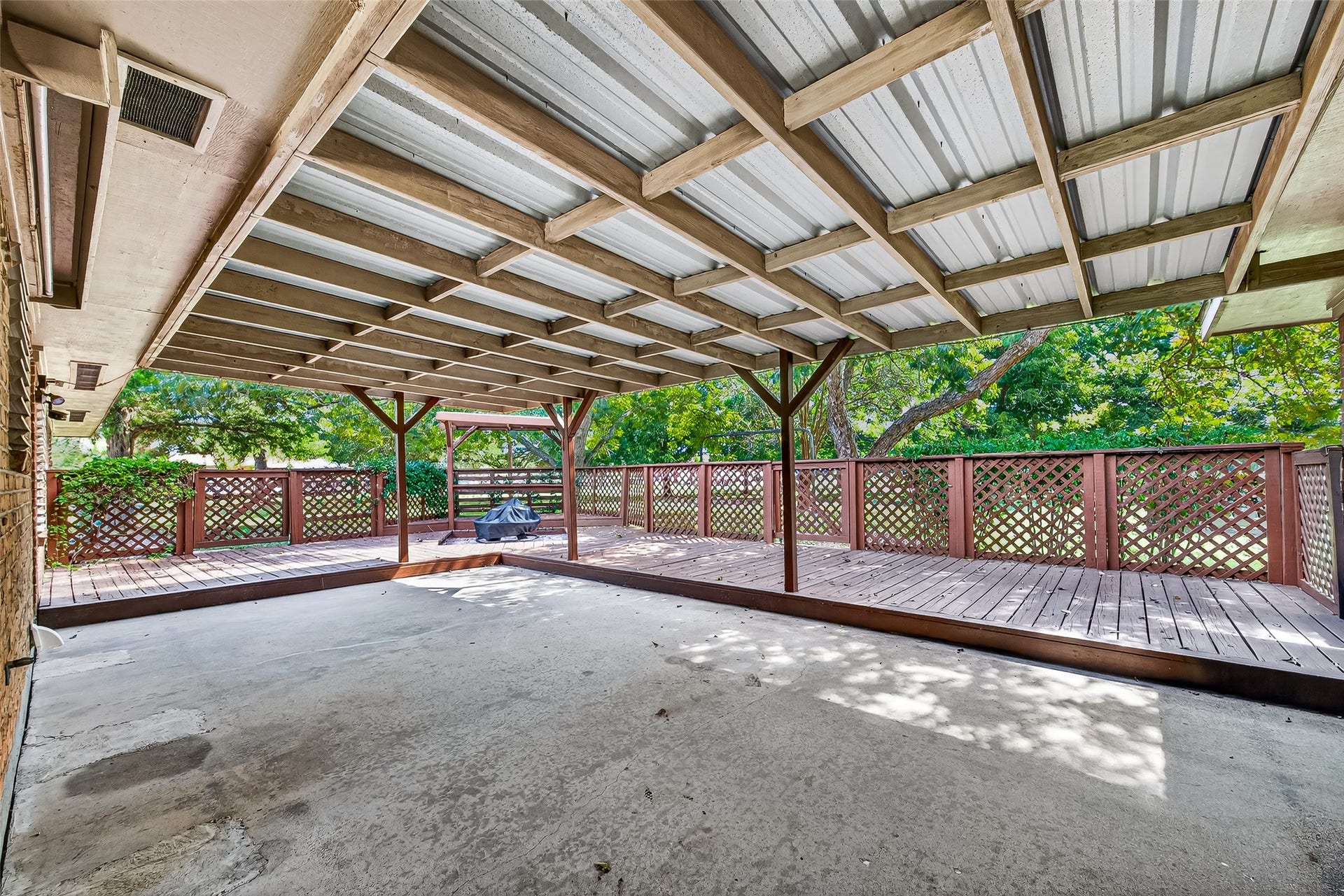 12021 7th 1/2 Street, Santa Fe, TX 77510 This photo showcases a spacious covered patio with a metal roof and wooden beams. It features a raised wooden deck with lattice railing, offering a private and shaded outdoor area surrounded by greenery, perfect for relaxation or entertaining.