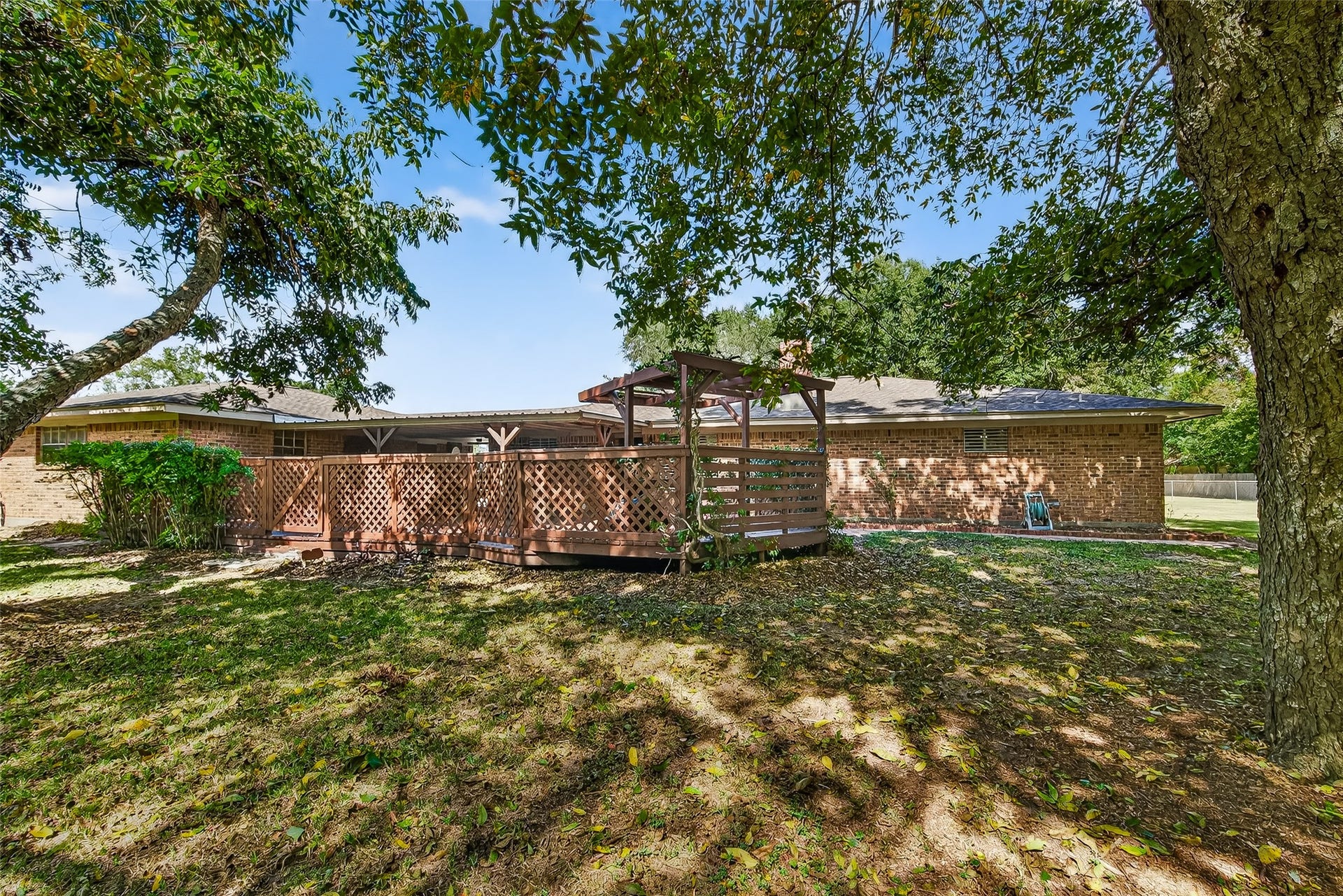 12021 7th 1/2 Street, Santa Fe, TX 77510 This photo showcases a backyard with a spacious wooden deck and lattice fencing, surrounded by mature trees providing shade. The brick exterior of the house complements the natural setting, creating a cozy and inviting outdoor space.