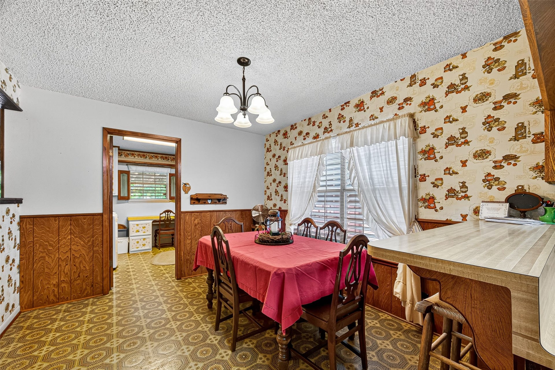 12021 7th 1/2 Street, Santa Fe, TX 77510 This cozy dining area features vintage wallpaper and wood paneling, with a bright window and overhead lighting. The space connects to a kitchen through a doorway, and includes a practical breakfast bar for additional seating.