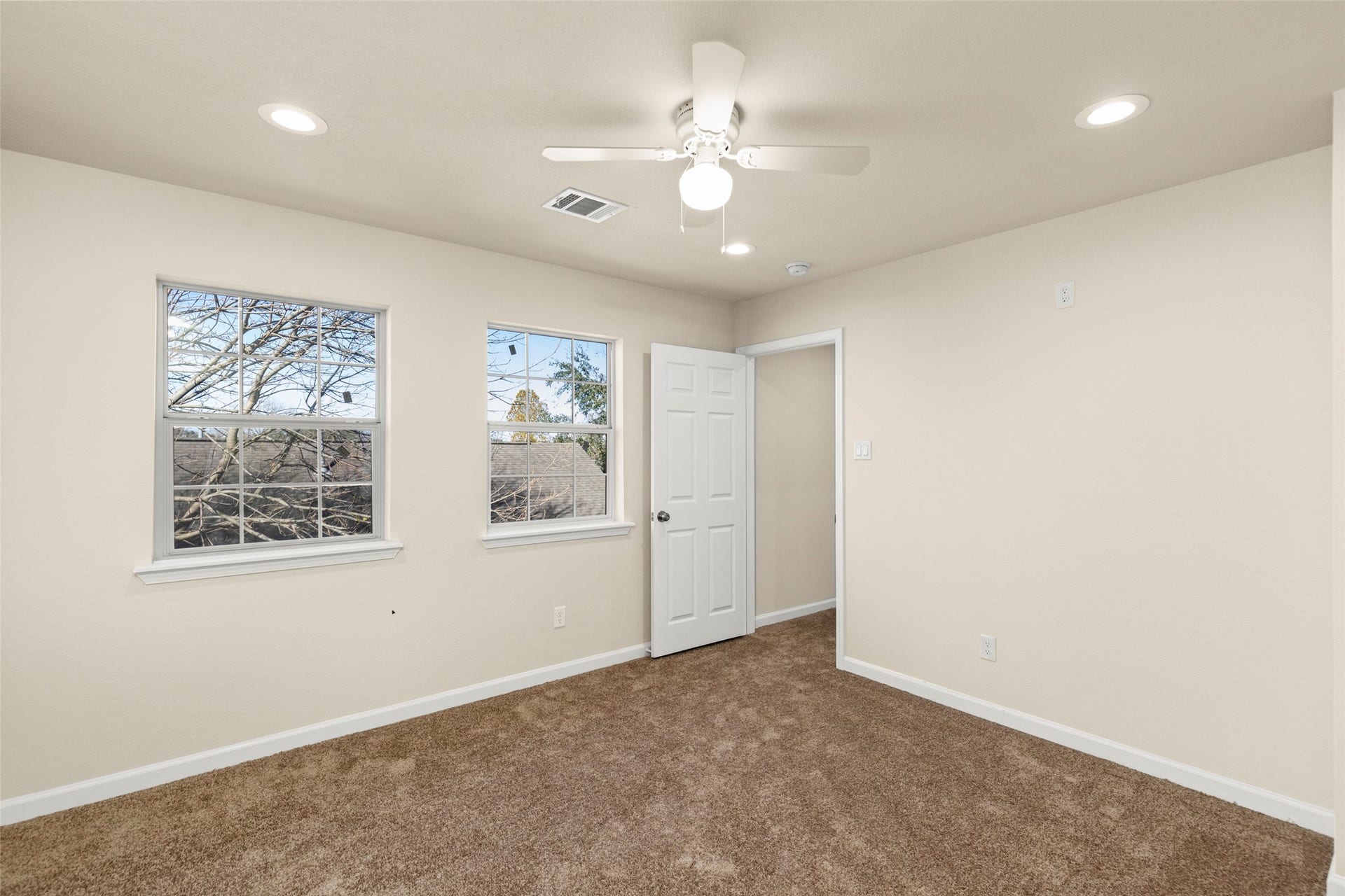 This hallway features neutral walls and carpet, leading to a bright room with windows. It includes access to a bathroom with a white vanity, offering a clean and inviting space.