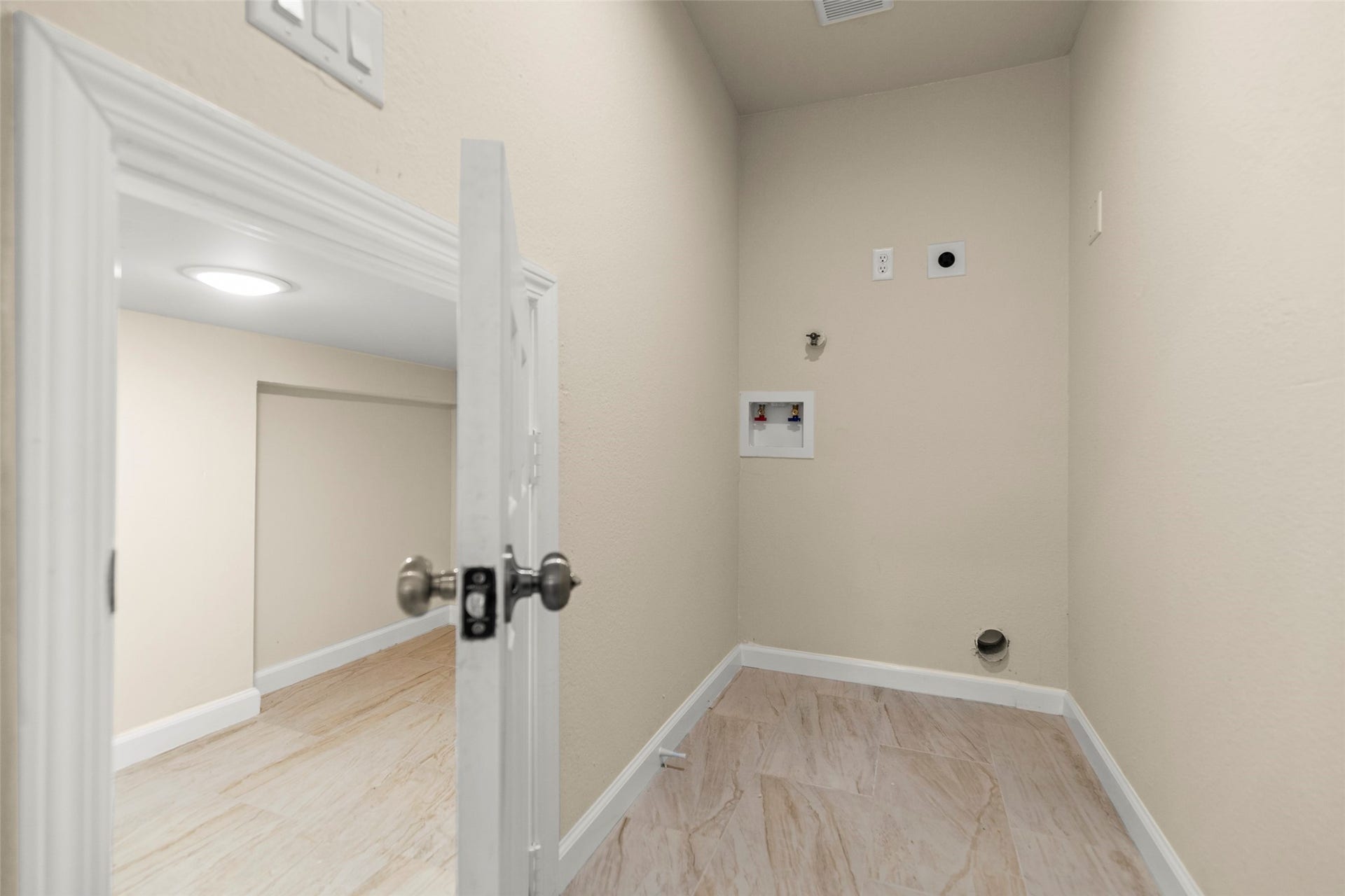 This photo shows a compact utility room with hook-ups for a washer and dryer. The room features light-colored walls and flooring, and there's a small adjacent space with its own door, possibly for storage or additional functionality.