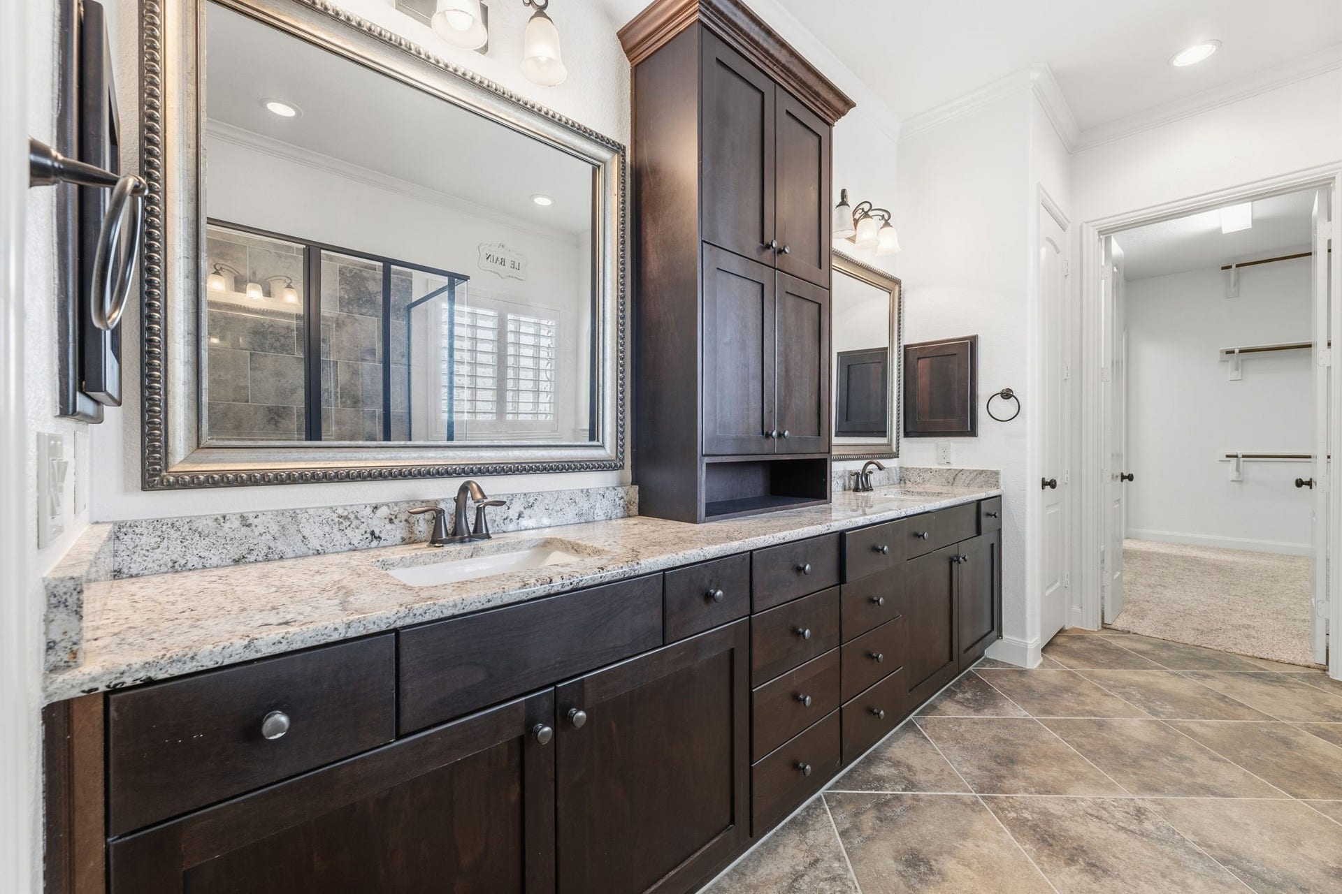 Primary bath featuring granite countertops, dark wood cabinetry, dual sinks, and a spacious layout that connects to a walk-in closet.