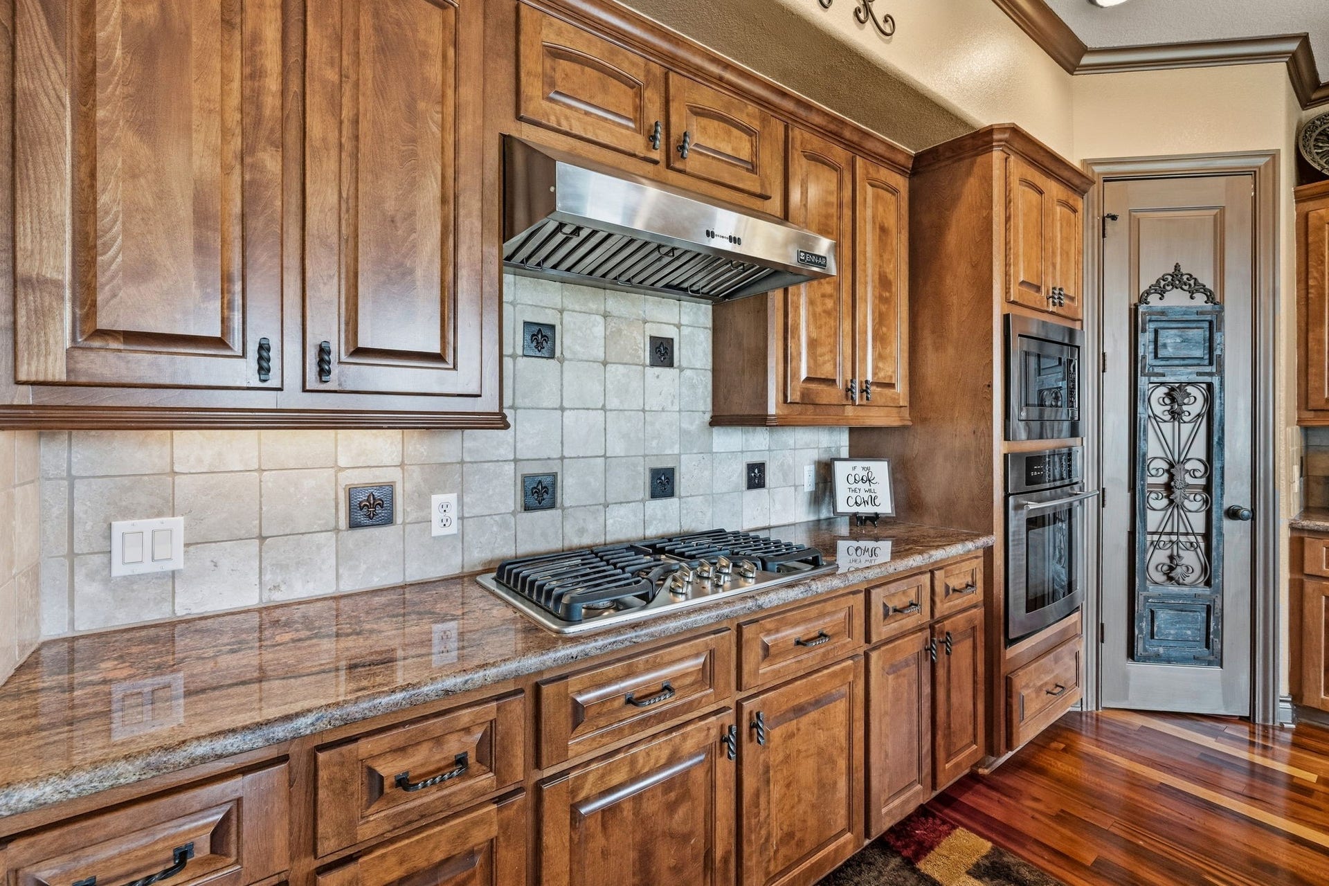 26 Half Moon, Hitchcock, TX 77563 Elegant kitchen featuring rich wood cabinetry, a sleek stainless steel range hood, and granite countertops. The tile backsplash and double ovens add a touch of sophistication, while a decorative pantry door completes the space.
